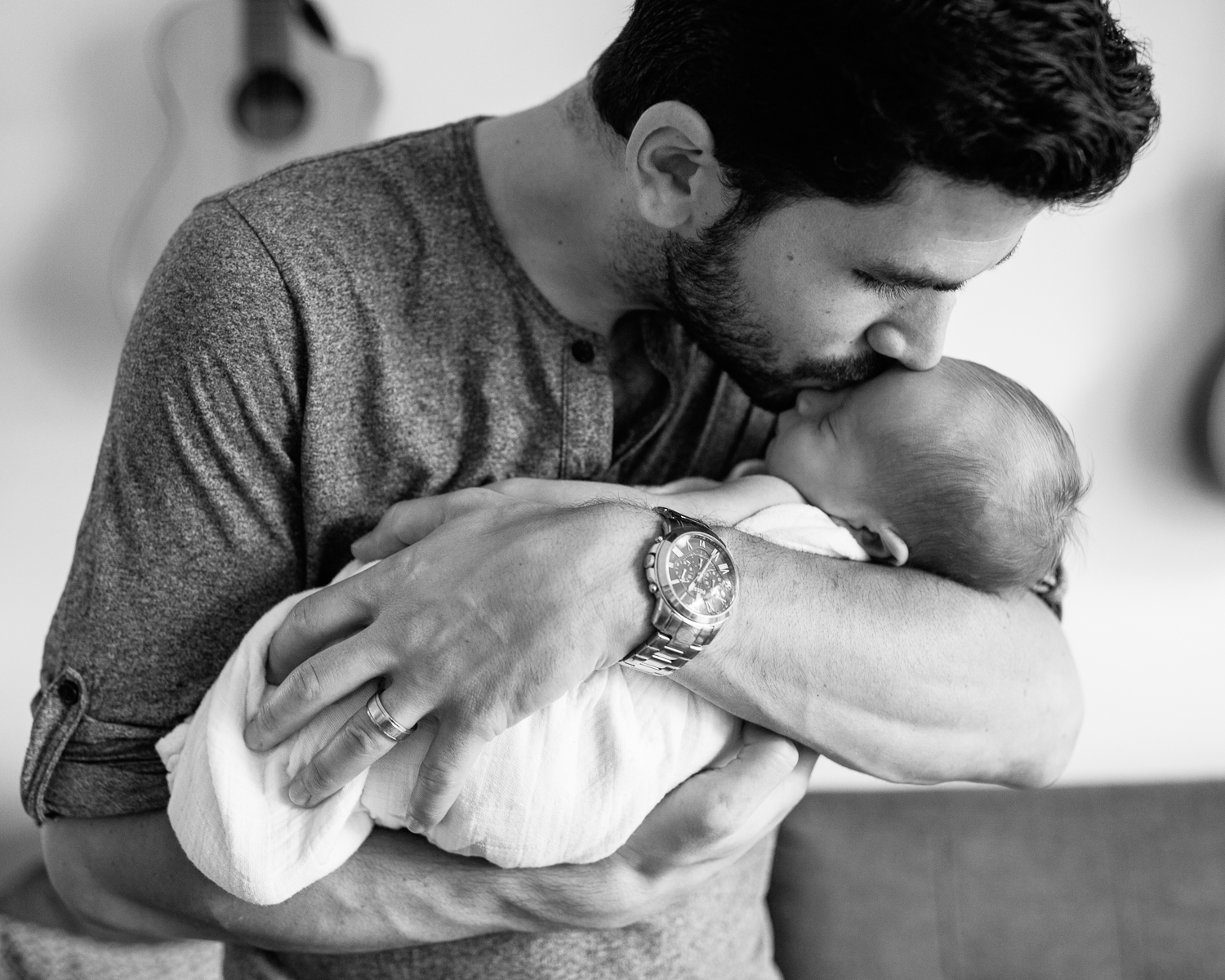 new dad standing in living room, holding 2 week old sleeping baby boy in white swaddle, kissing son on forehead - York Region Lifestyle Photography