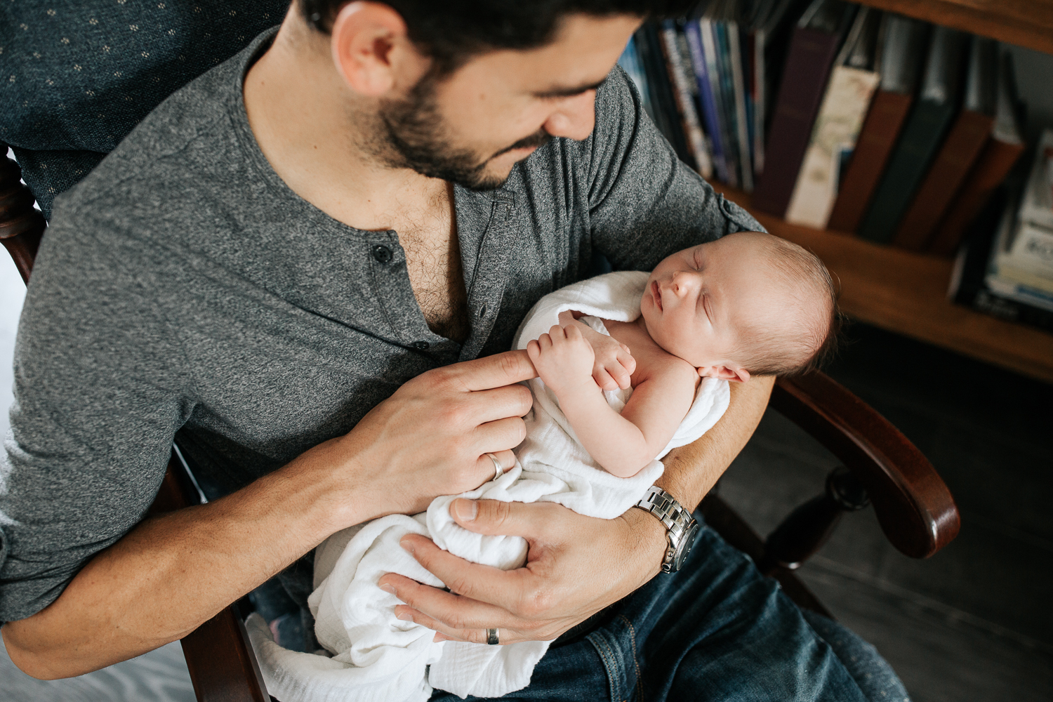 new dad sitting in glider, holding 2 week old sleeping baby boy in white swaddle, son holding father's finger in his hand - GTA Lifestyle Photography