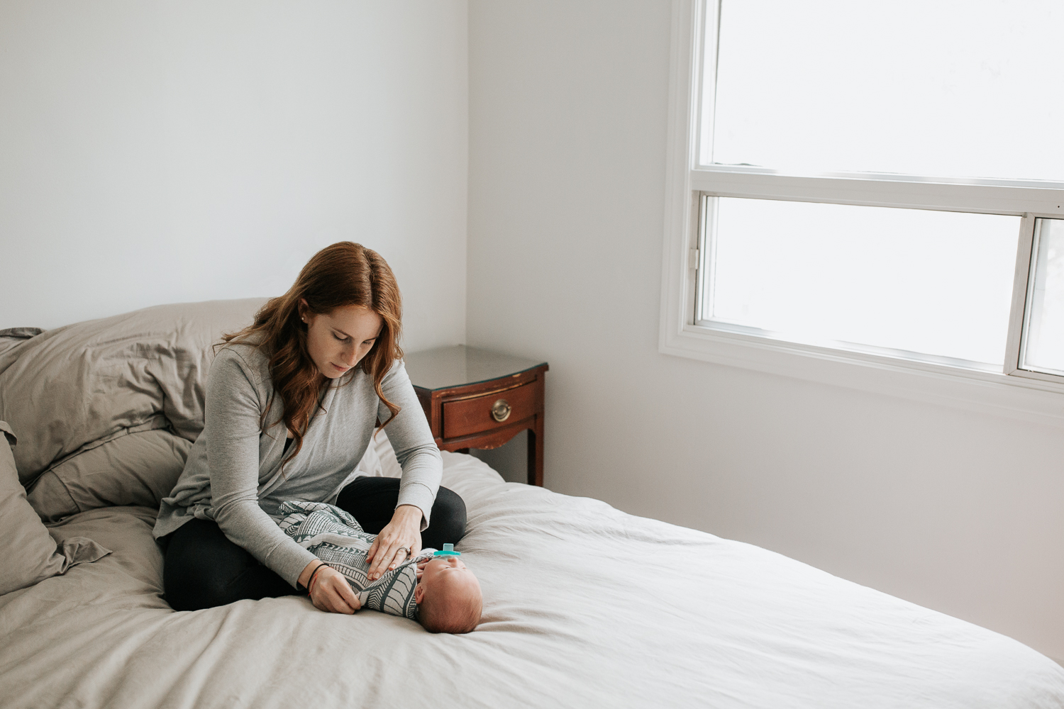 new mother with long red hair sitting cross legged on master bed swaddling 2 week old baby boy - Barrie Lifestyle Photography