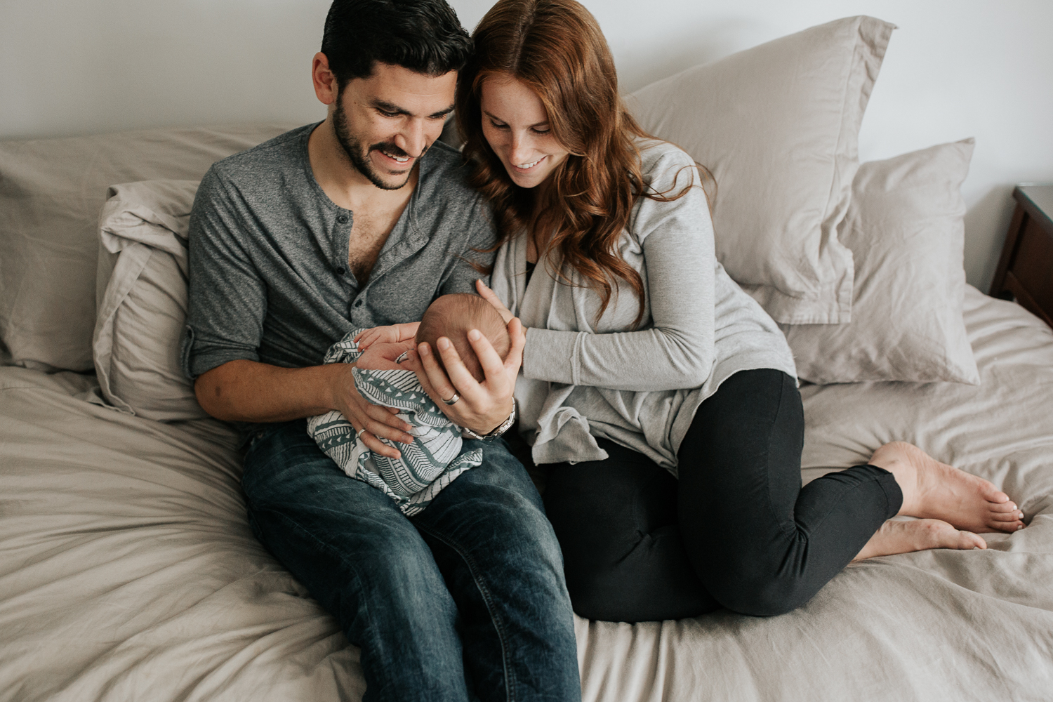 new parents sitting on master bed, dad holding 2 week old baby boy, mom snuggled next to them, smiling at son - Stouffville Lifestyle Photography