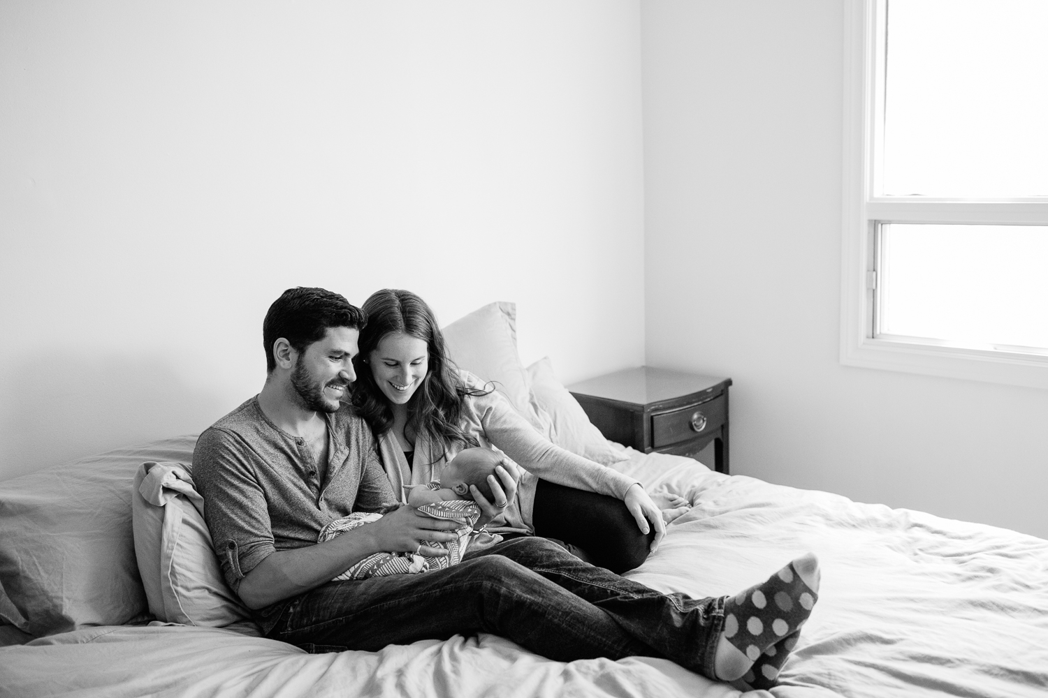 new parents sitting on master bed, dad holding 2 week old baby boy, mom snuggled next to them, smiling at son - Markham Lifestyle Photography