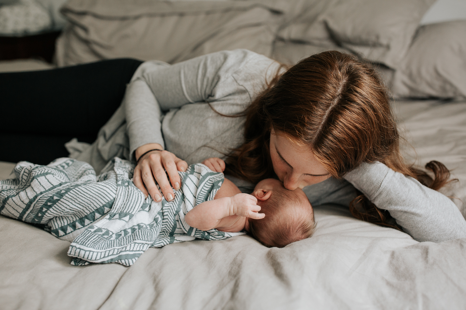 new mom with long red hair lying on bed next to 2 week old baby boy in geometric swaddle, kissing him on forehead - GTA Lifestyle Photography