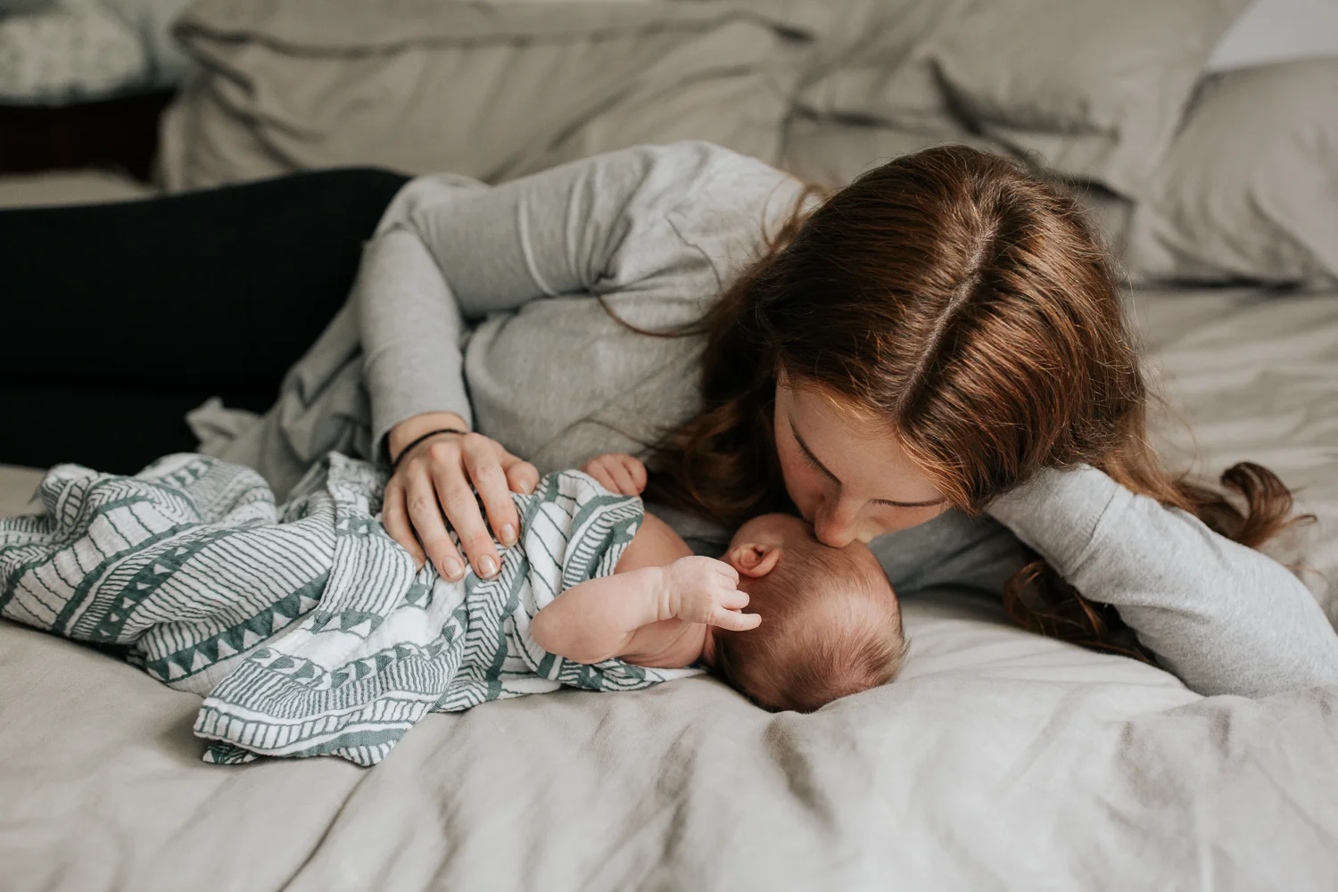 new mom with long red hair lying on bed next to 2 week old baby boy in geometric swaddle, kissing him on forehead - GTA Lifestyle Photography