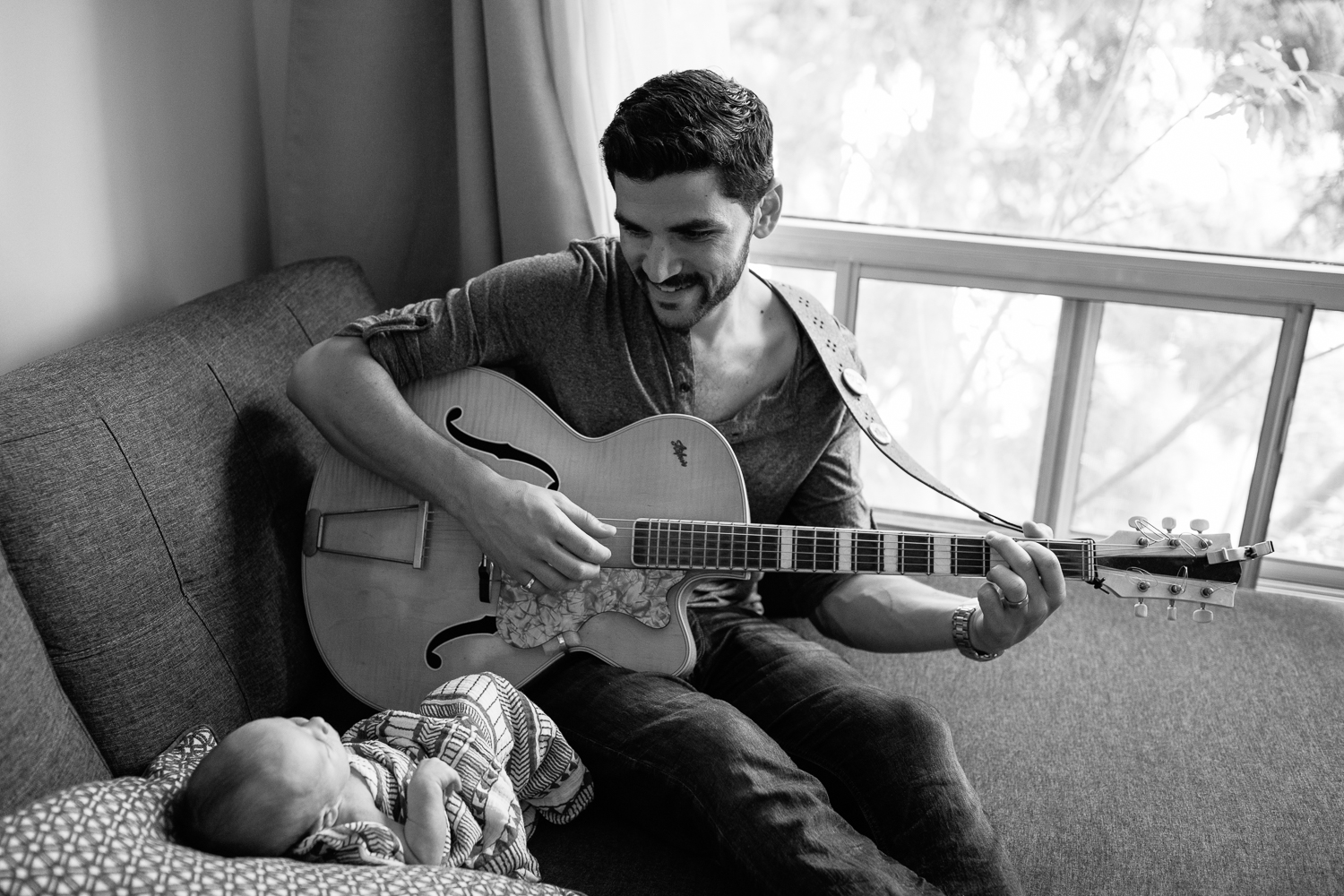 new dad sitting on living room couch playing guitar and singing to 2 week old baby boy lying against cushions - York Region Lifestyle Photography