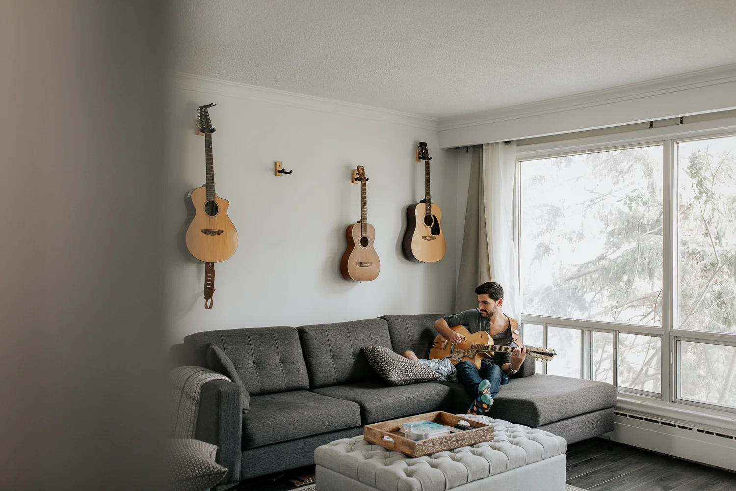 new dad sitting on living room couch playing guitar and singing to 2 week old baby boy lying against cushions - Newmarket In-Home Photography