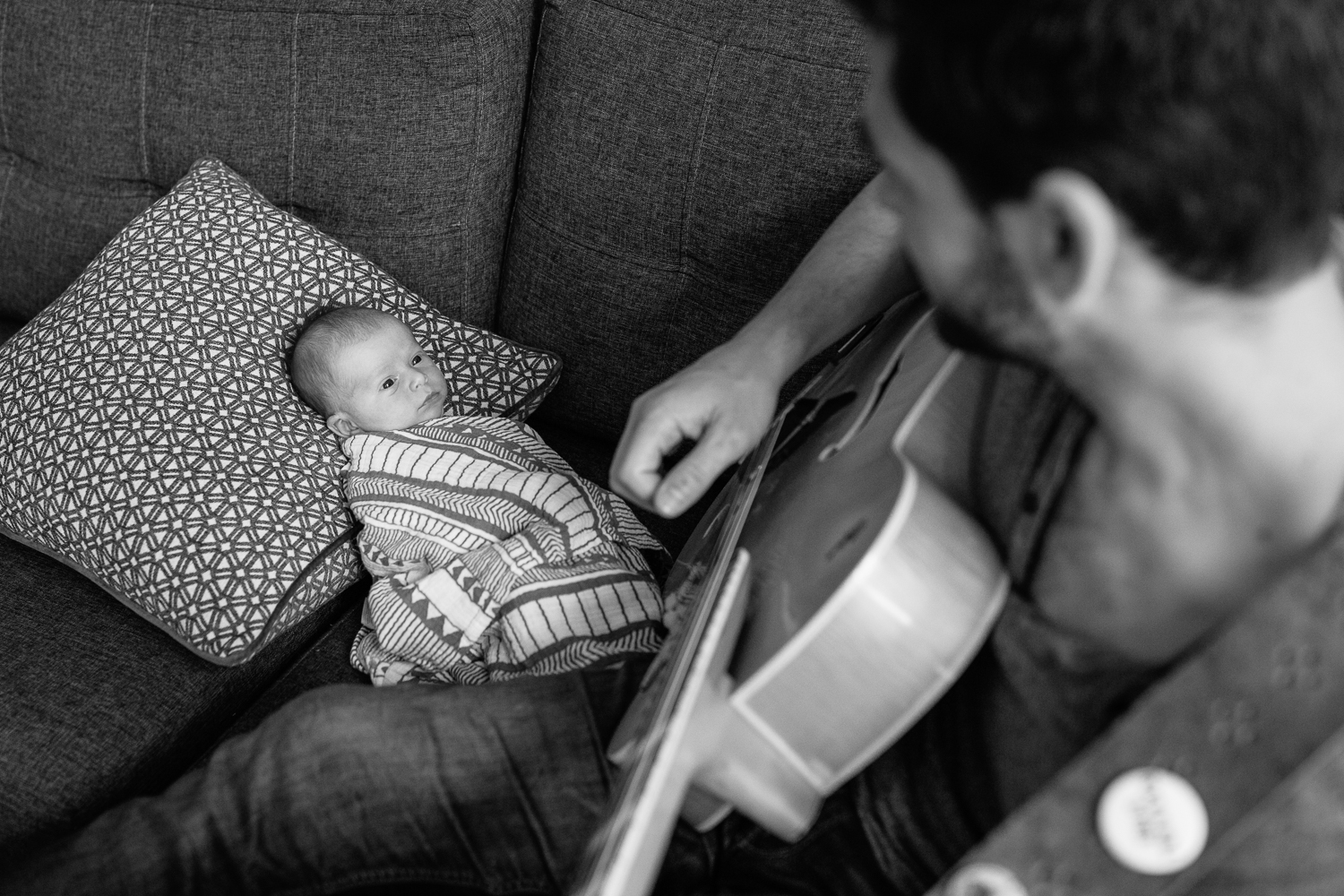 new dad sitting on living room couch playing guitar and singing to 2 week old baby boy lying against cushions watching father - Stouffville In-Home Photography