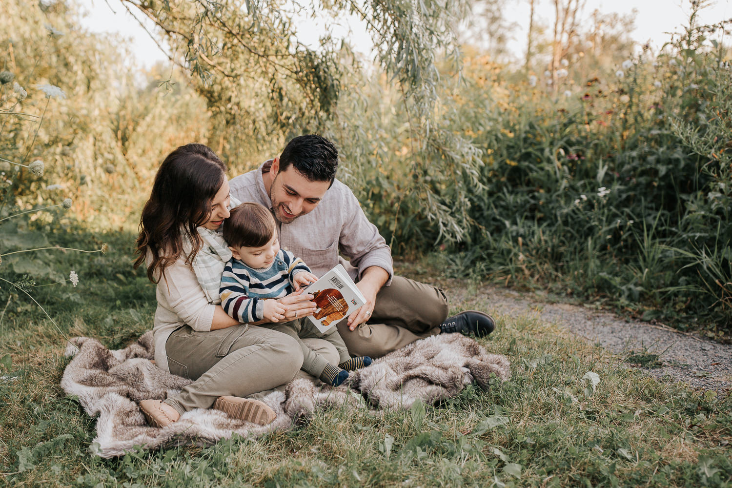 family of 3 sitting on fur blanket under willow tree at sunset, 1 year old baby boy sitting on mom's lap reading story, dad next to them smiling down at son - Newmarket Lifestyle Photography