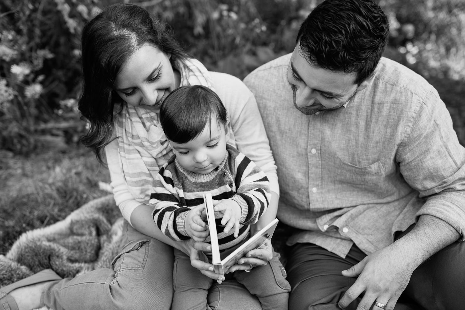 Markham Lifestyle Photographyfamily of 3 sitting on fur blanket under willow tree at sunset, 1 year old baby boy sitting on mom's lap reading story, dad next to them smiling down at son - 