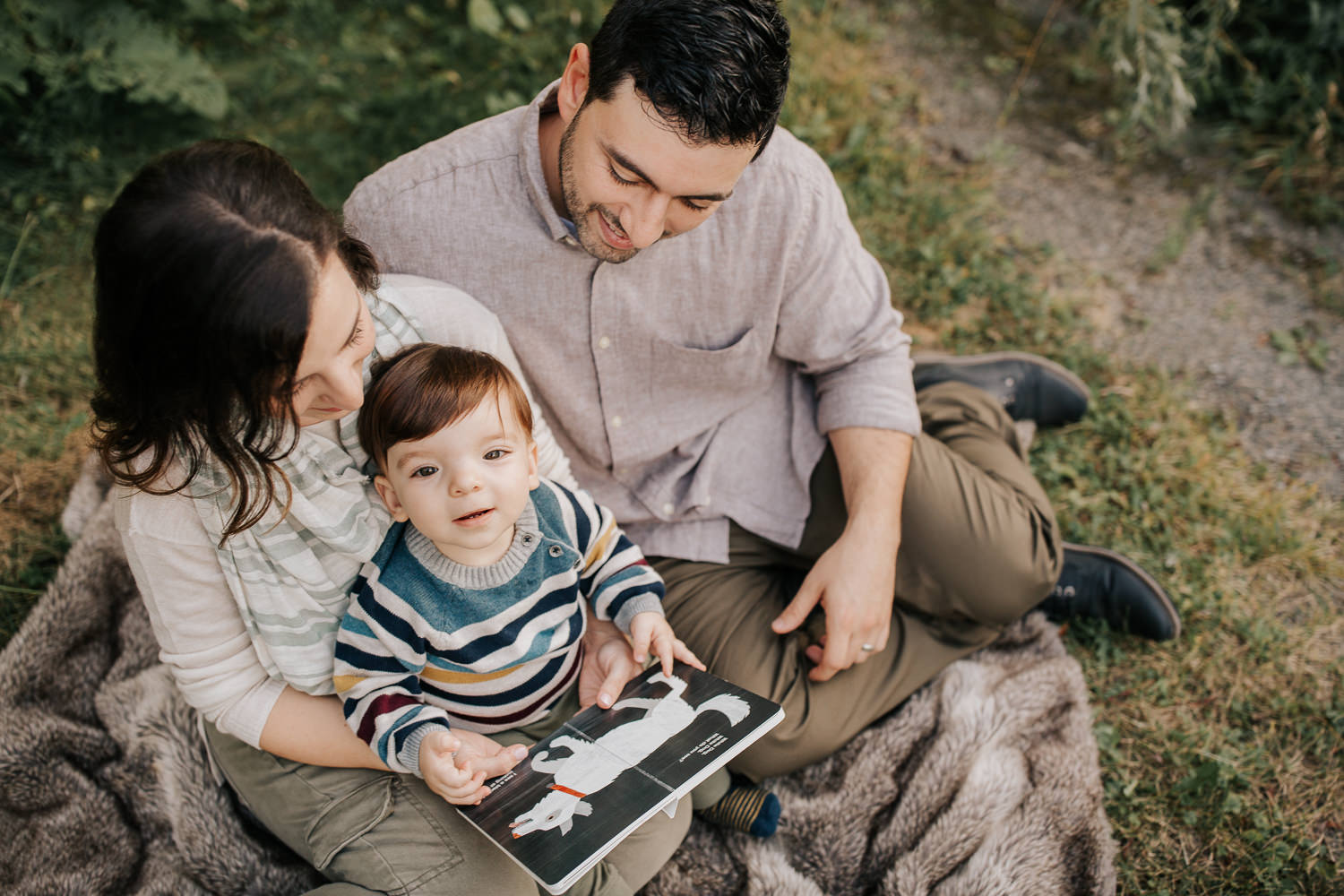 family of 3 sitting on fur blanket under willow tree at sunset, 1 year old baby boy sitting on mom's lap, looking up at camera, dad next to them smiling down at son - GTA Lifestyle Photography