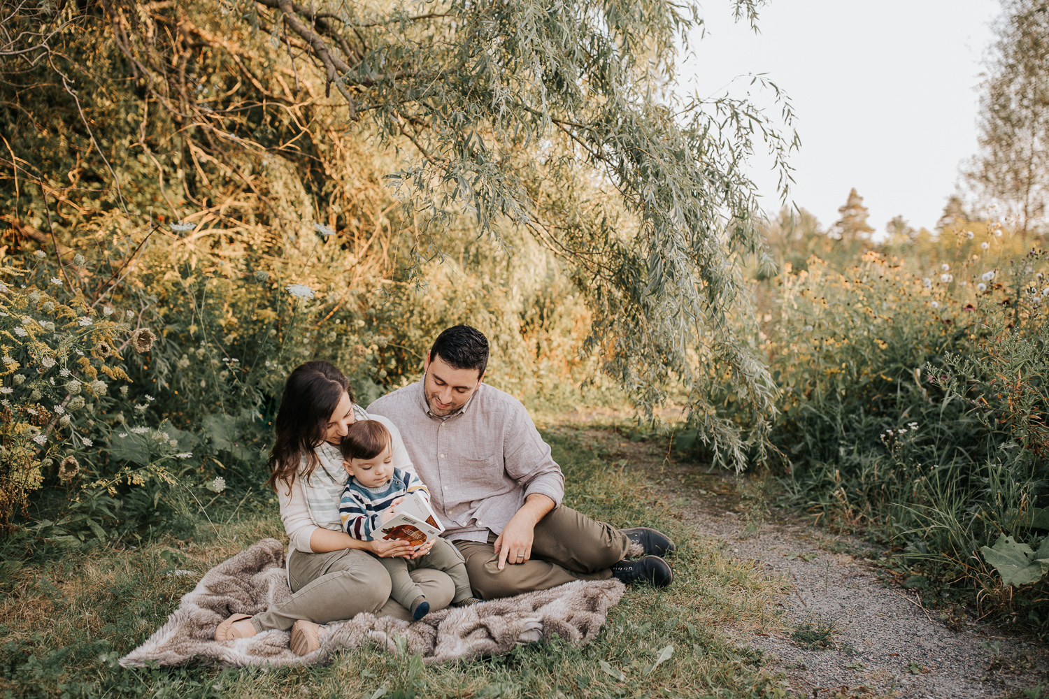 family of 3 sitting on fur blanket under willow tree at sunset, 1 year old baby boy sitting on mom's lap reading story, dad next to them smiling down at son - Barrie Lifestyle Photography
