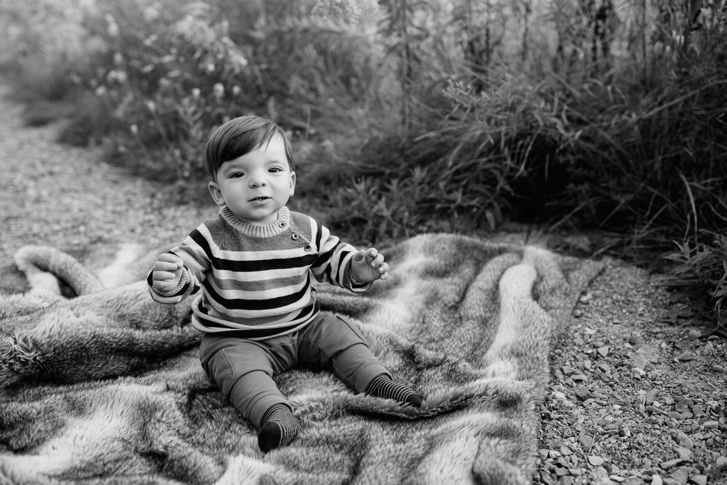1 year old boy with dark hair and eyes wearing striped sweater sitting on blanket in outdoor path looking at camera - York Region Golden Hour Photography