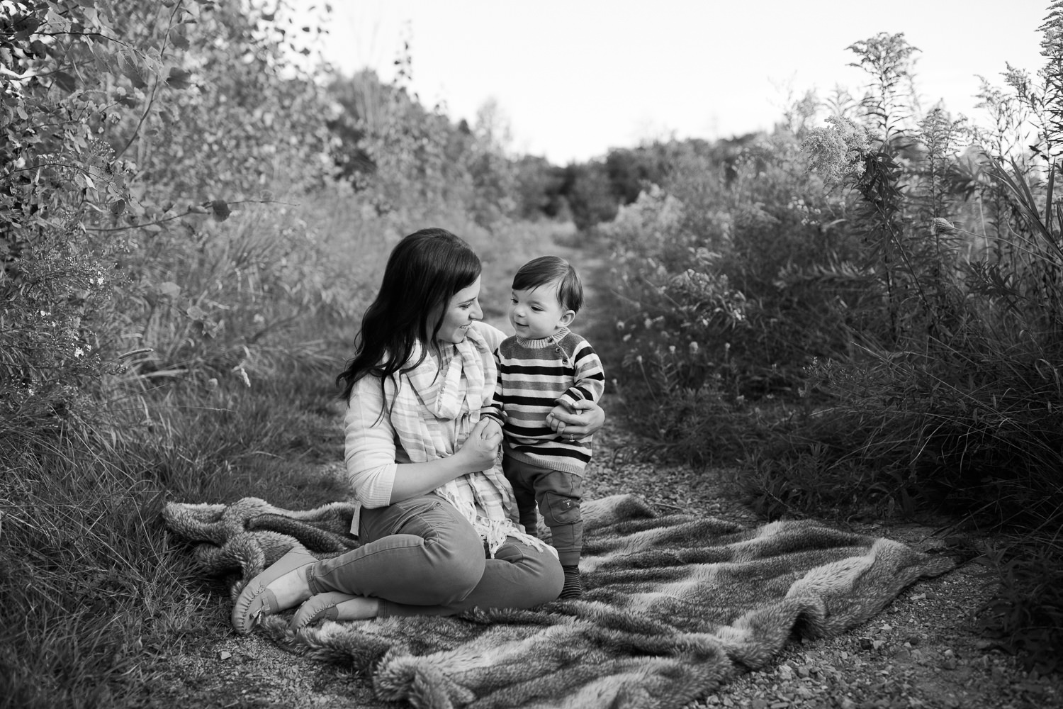 mother sitting on fur blanket on outdoor grassy path, 1 year old baby boy standing next to her, mom and son smiling at each other - GTA Golden Hour Photography