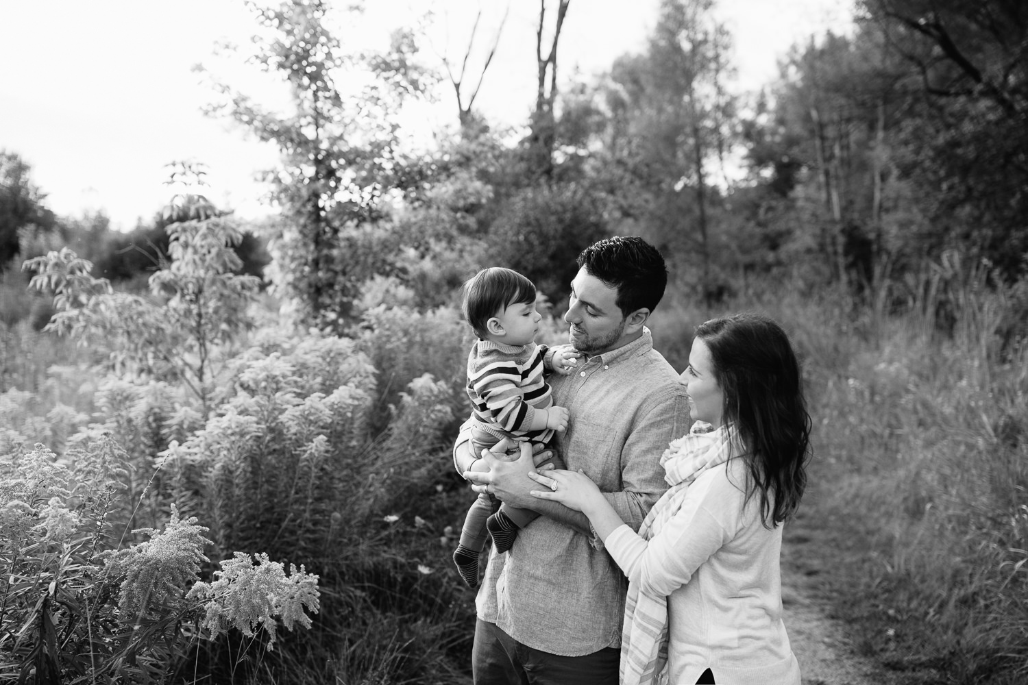 family of 3 standing on path in grassy field, dad holding 1 year old baby boy, mom standing behind husband, hand on his arm, looking at son - GTA Golden Hour Photos