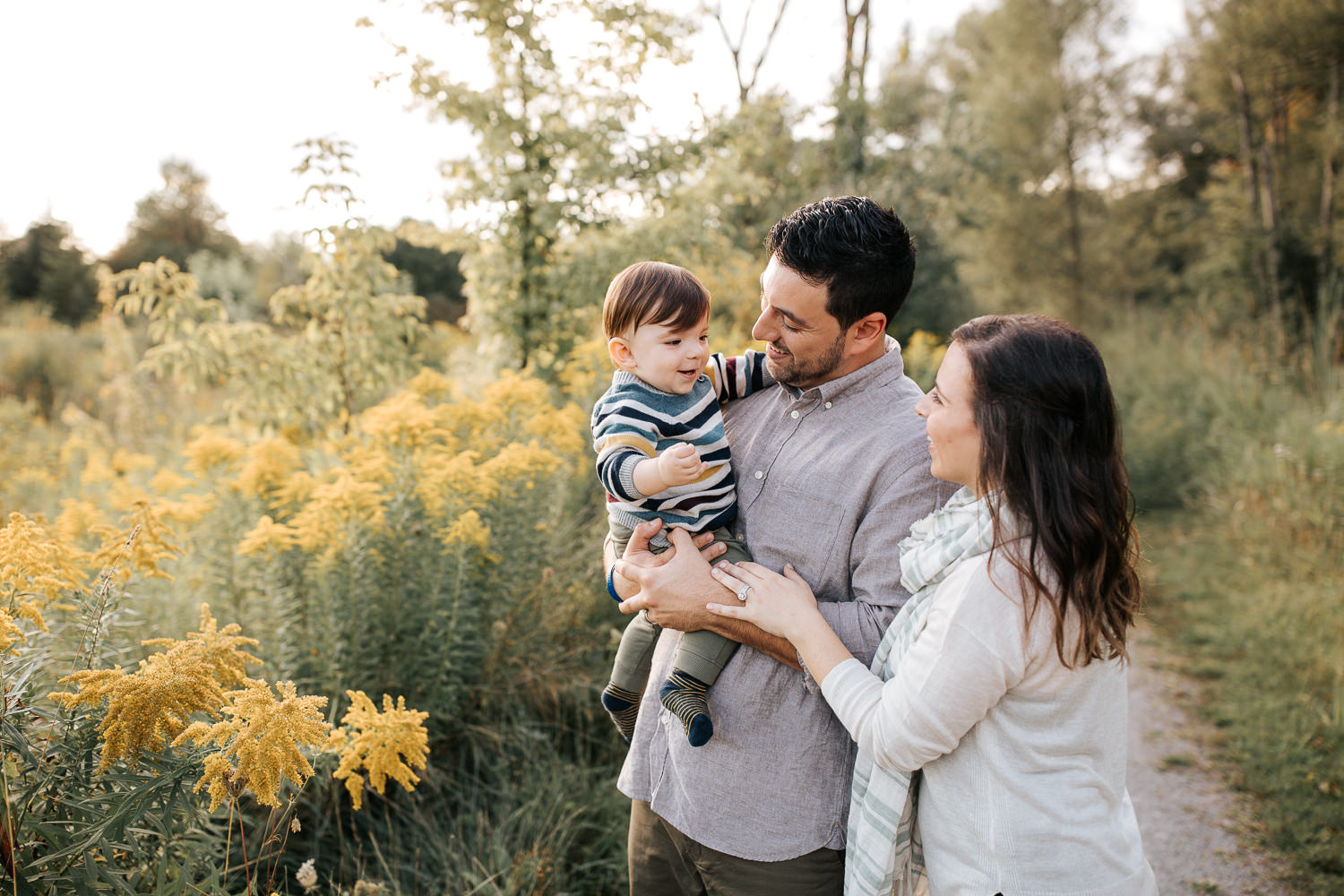 family of 3 standing on path in golden field, dad holding 1 year old baby boy, mom standing behind husband, hand on his arm, looking at son - Stouffville Lifestyle Photos