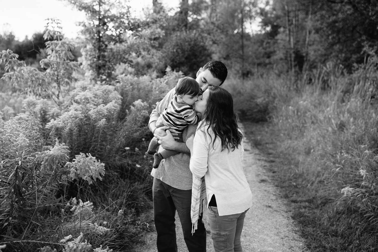 family of 3 standing on path in grassy field, dad holding 1 year old baby boy, mom standing next to husband, leaning over to kiss son - Markham Lifestyle Photos