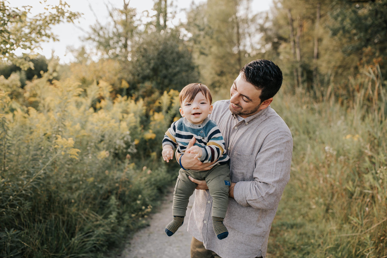 dad standing in golden field holding 1 year old baby boy, son smiling at camera, father smiling at him - York Region Lifestyle Photos