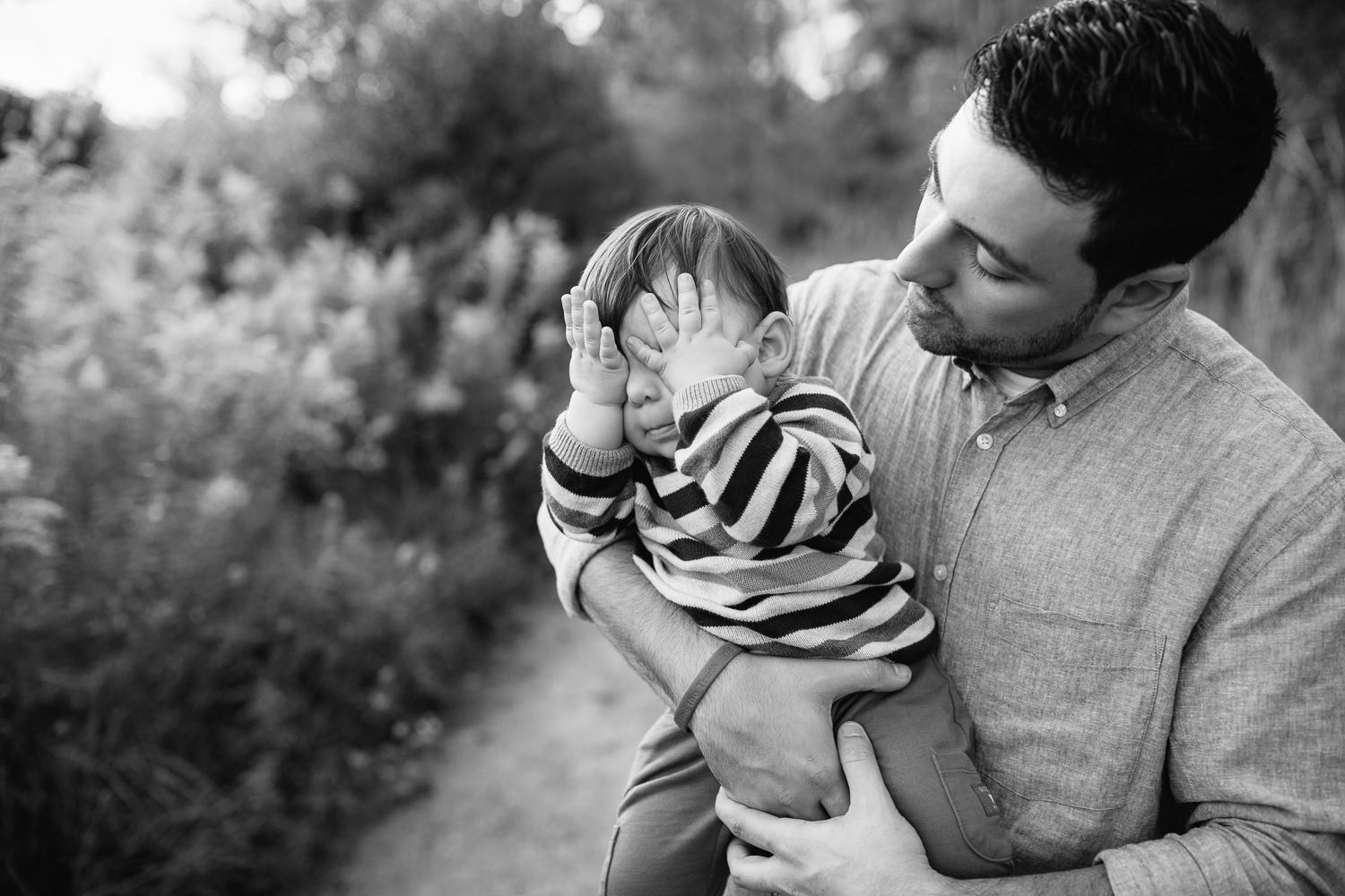 dad standing in golden field holding 1 year old baby boy, child with hands over eyes playing peek-a-boo, father smiling at son - Newmarket Lifestyle Photography