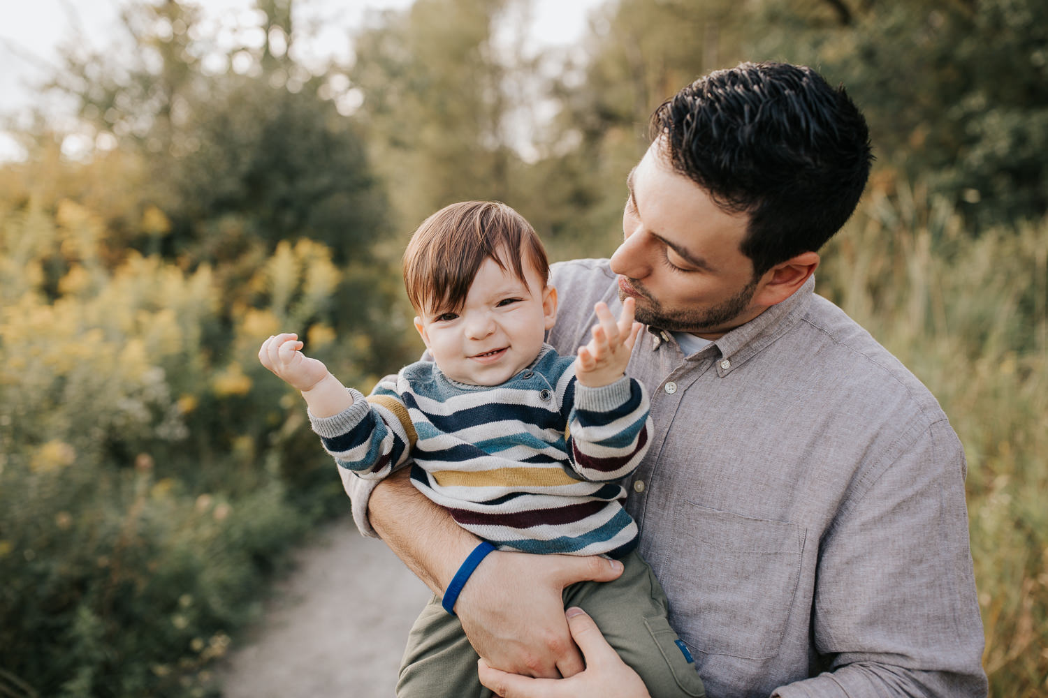 dad standing in golden field holding 1 year old baby boy, child playing peek-a-boo, father smiling at son - Stouffville Lifestyle Photography