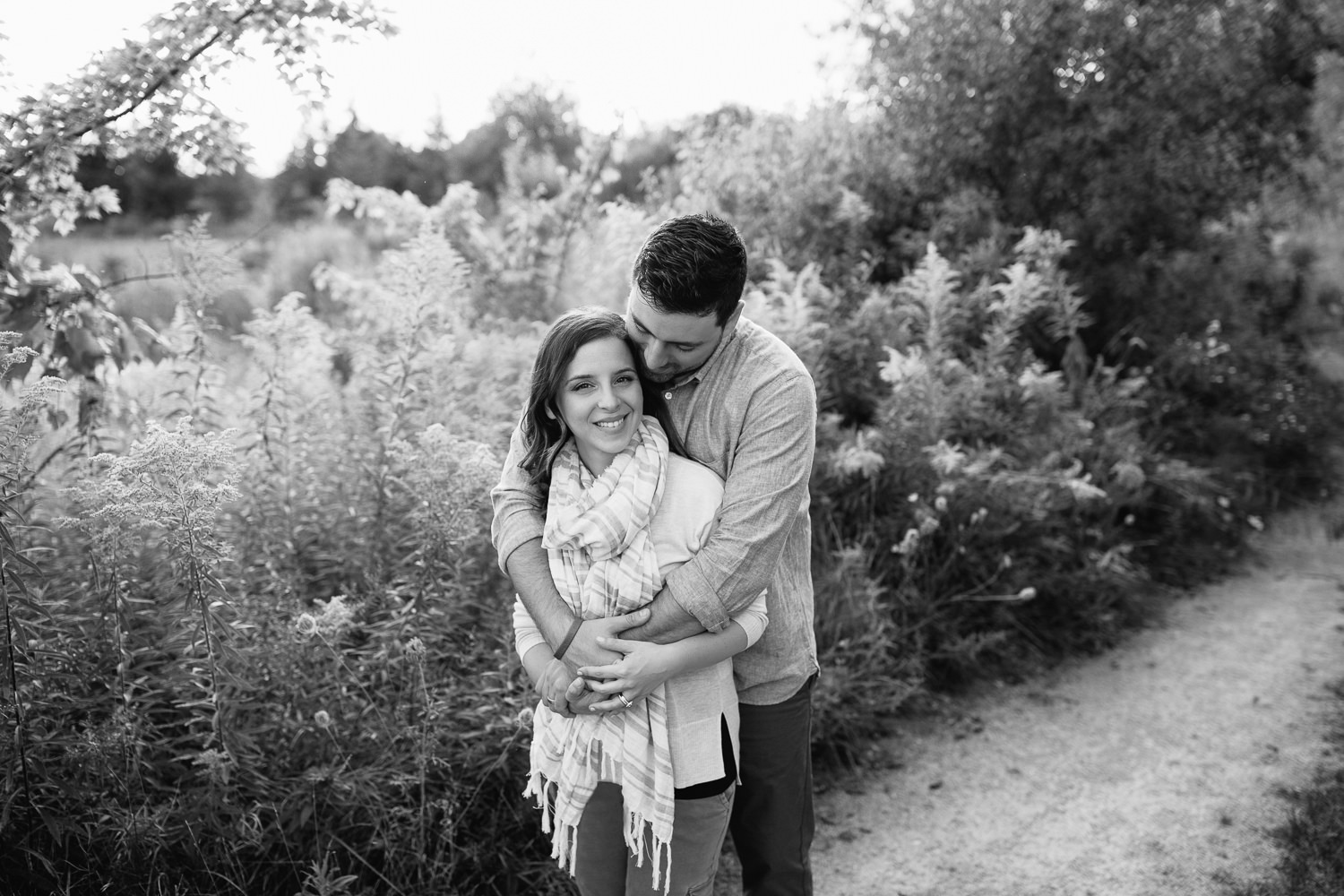 couple standing on path at sunset, sun setting behind them, husband standing behind wife embracing her, woman smiling at camera - Markham Lifestyle Photography