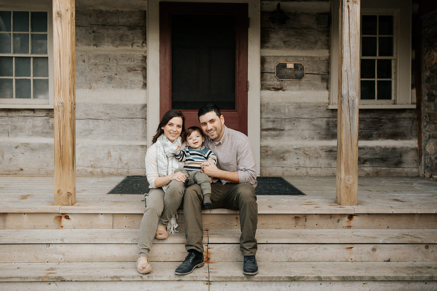 family of 3 sitting on front porch steps of historic log cabin, son on dad's lap, mom next to them, smiling at camera - Stouffville Outdoor Photography