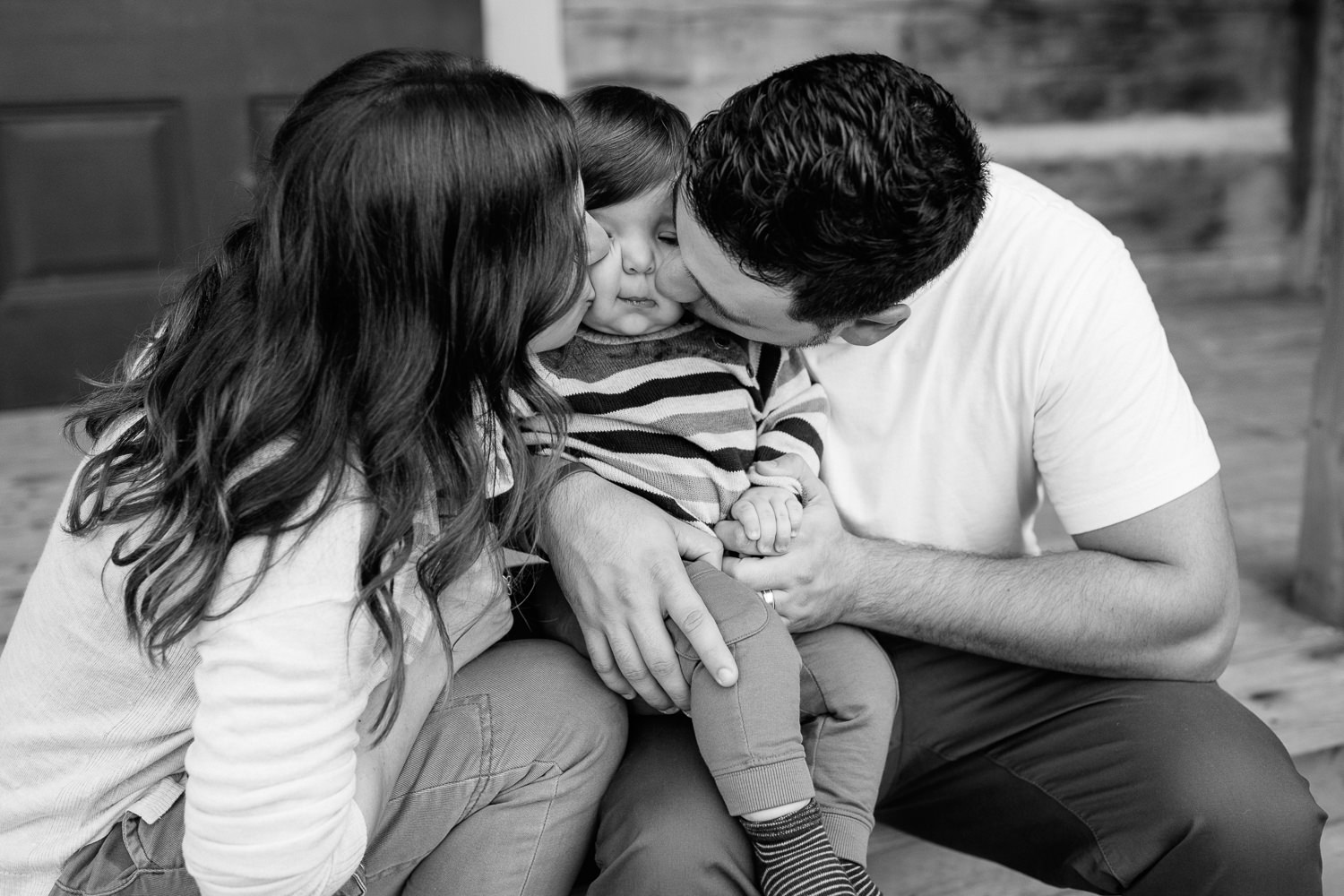 family of 3 sitting on front porch steps of historic log cabin, son between mom and dad, parents kissing child on cheeks - York Region Outdoor Photography