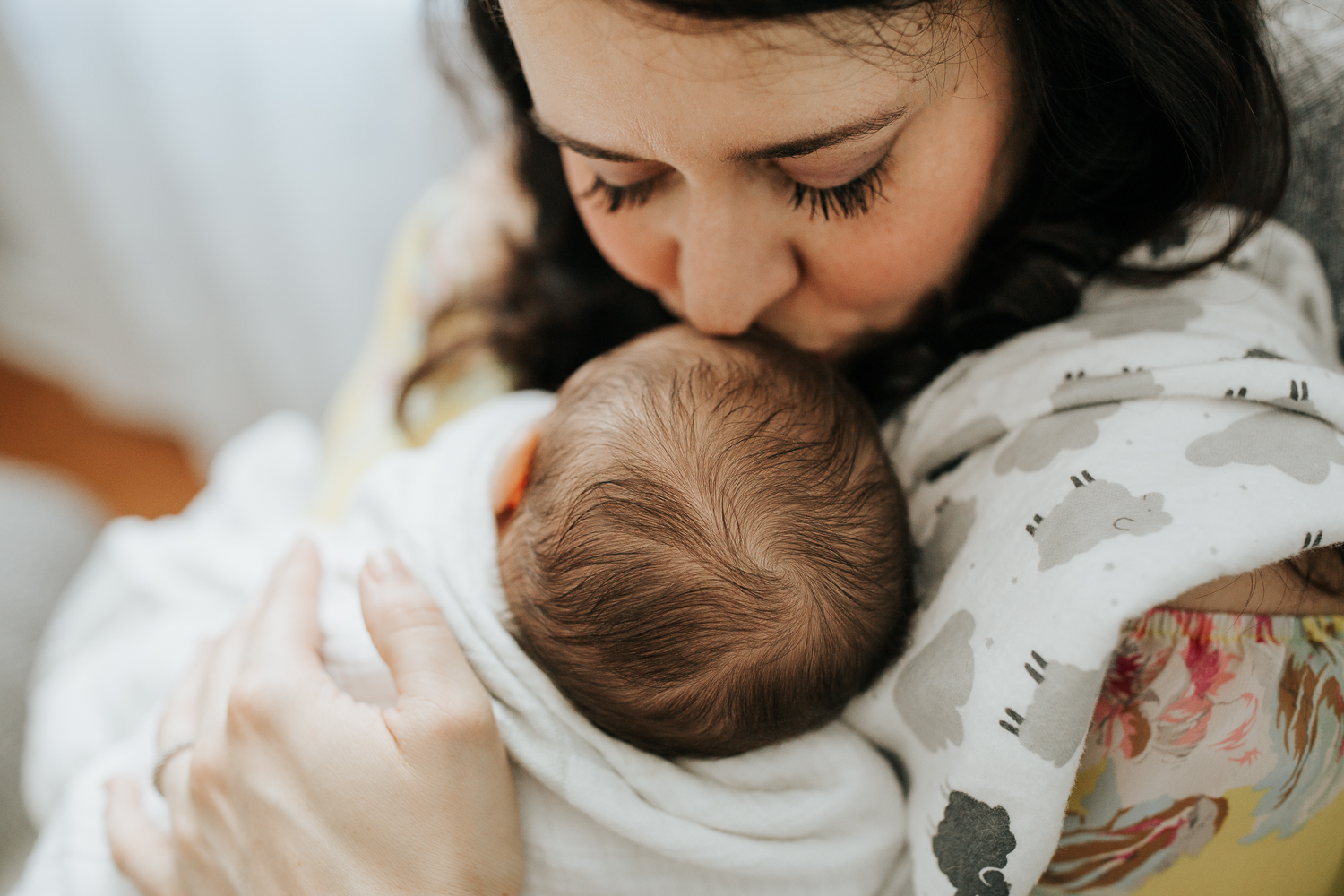 new mother sitting in chair, 2 week old baby boy swaddled and lying on her chest, mother kissing son's forehead - York Region Lifestyle Photography