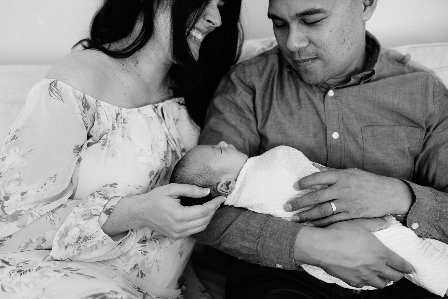 new parents sitting on couch, 3 week old baby boy in father's arms, mother smiling at husband, black and white - York Region Lifestyle Photography