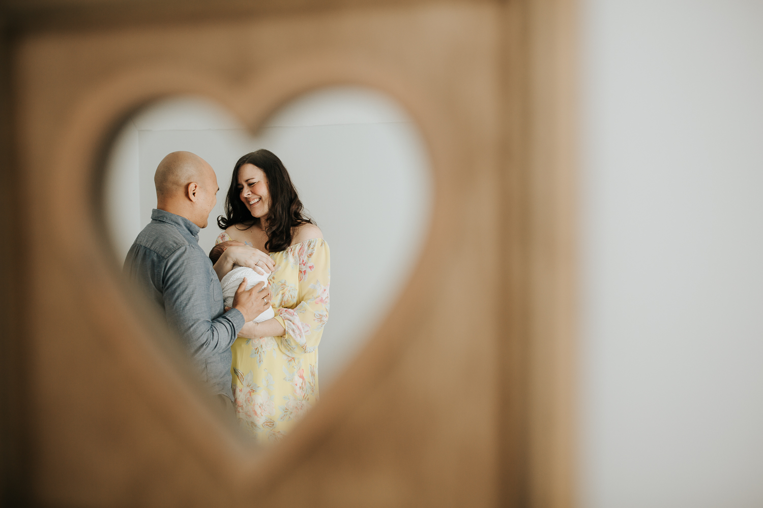 new parents framed in heart mirror, mother holding sleeping 3 week old baby boy to her chest, smiling at husband, dad's hand on son - GTA Lifestyle Photos