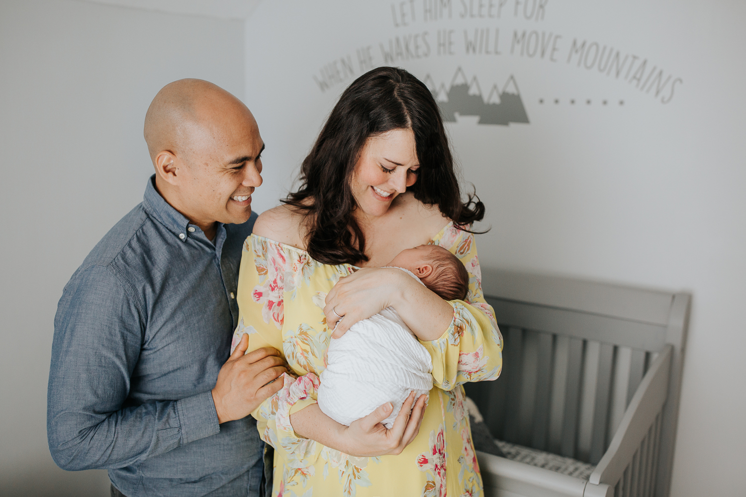 new parents standing in front of crib, mom holding 3 week old sleeping, swaddled baby boy to her chest, husband standing next to wife, hand on her arm - GTA Lifestyle Photos