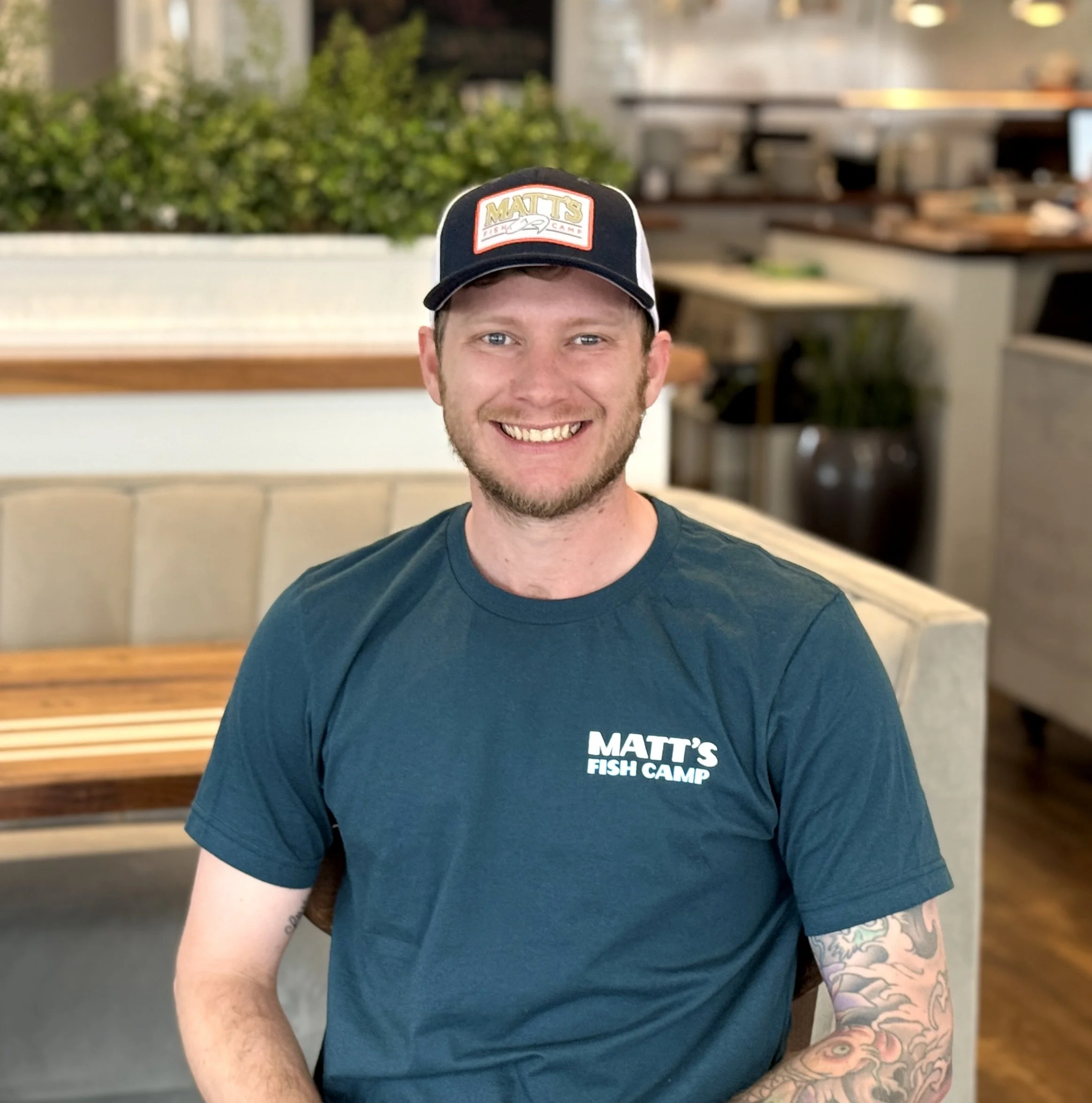 A man with tattoos on his right arm wearing a navy blue t-shirt with the text "Matt's Fish Camp" and a navy and white baseball cap with the same logo, smiling at the camera in a restaurant or cafe setting.