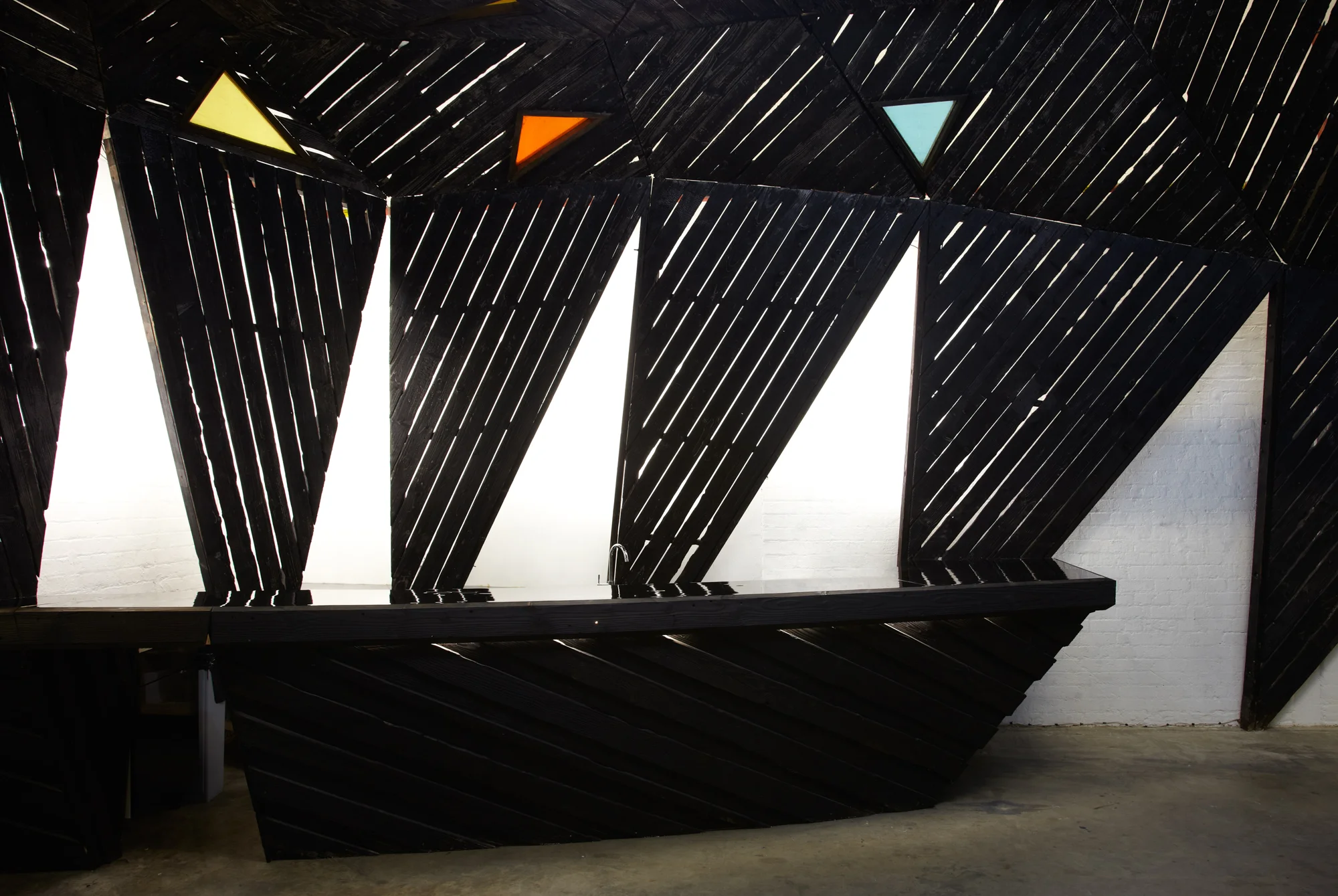 ‘Black Pleasure’ is the new public space within Eastside Projects, by artists Heather & Ivan Morison, formed from the meeting and merging of two of the artists’ long term, large scale artworks – ‘Pleasure Island’ and ‘Black Cloud’. ‘Black Pleasure’ provides new opportunities for the life of the gallery at Eastside Projects as it opens up a new communal space for performing, sharing, talking, eating and drinking. The artwork is a useful space as much as it in turn reflects upon the prophetic visions of twentieth century science fiction writers, the urgent contemporary concerns of the Arts & Ecology movement, and the real world application of collectivist ideals within controlled environments. ‘Pleasure Island’ was the first artwork constructed as part of Eastside Projects in 2008, functioning as both central office and kitchen for the gallery. Over the last five years ‘Pleasure Island’ has acted as a beacon of the myth of a gallery that is an artwork. The Morison’s much loved structure was built from harvested red wood trees from the artists’ own wood in Wales and its form was based on a Peruvian fools-gold crystal gifted to them. Originally commissioned for the Wales Pavilion at the Venice Biennale in 2007 ‘Pleasure Island’ was adapted for Eastside Projects as a long term commitment to exploring the nature of artworks within the space. ‘Pleasure Island’ was also the name of the artists’ first puppet play within the ‘office’ on the launch of the gallery in 2008 and this has been followed by four further puppet performances,culminating in ‘Puppet Show’ earlier this year. ‘The Black Cloud’ was proposed by Heather & Ivan Morison as a shelter for a future apocalyptic world scorched black by the unrelenting sun. A remarkable pavilion-like structure commissioned by Situations and first erected in Victoria Park, Bristol in 2009, ‘The Black Cloud’ acts as a public artwork, shelter and meeting place, and was mostrecently found ‘floating’ in the grounds of Hepworth Wakefield in Yorkshire. The dark structure was first erected through community 'barn raising' and was informed by a number of architectures from across the world responding to climatic scenarios or emergencies,such as the Hallig houses of Northern Germany built to withstand regular flooding, which echoes Eastside Projects’ own position between Heath Mill Lane and Floodgate Street named for their proximity to Birmingham’s main river, the Rea. ‘The Black Cloud’s starting point though was the ‘shabono’, a circular structure built by the Yanomamo South American Indians from the Amazon, made up of successive rings of timber triangular frames, bolted together to create a strong curving roof with an open centre, reflecting the communal space of the shabono. Constructed from trees from the same wood as ‘Pleasure Island’, the timber has been scorched using a Japanese technique, called Yakisugi treatment, to create a dark, protective shield. Between the current moment and a potential future catastrophe ‘The Black Cloud’ has moved on, and ‘Pleasure Island’ has not outstayed its welcome, nor has either structure lingered long enough to become commonplace. As we at Eastside Projects grow and change, and our needs and desires are honed, ‘Black Pleasure’ emerges out of the ashes of two artworks to satisfy our demands. A new myth emerges. Gavin Wade, Director, Eastside Projects, Birmingham