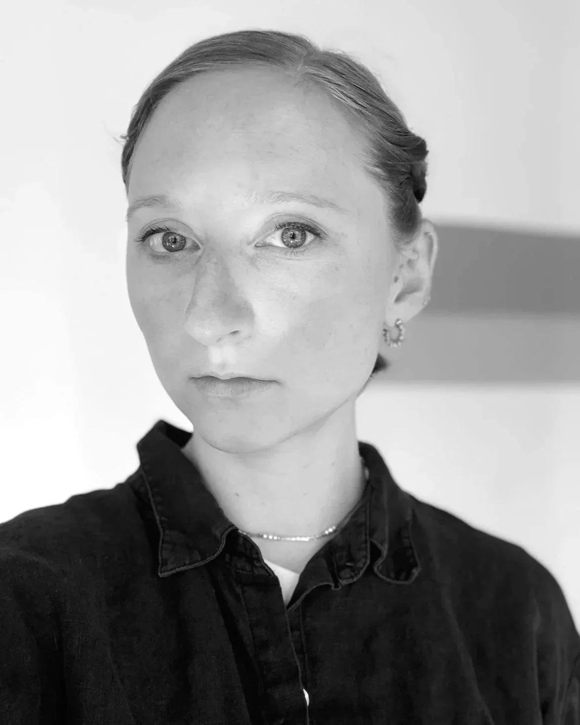 Black and white portrait of a femme presenting with golden blonde hair bulled back. They're wearing a black collared shirt, earrings, and a silver necklace with small pearls