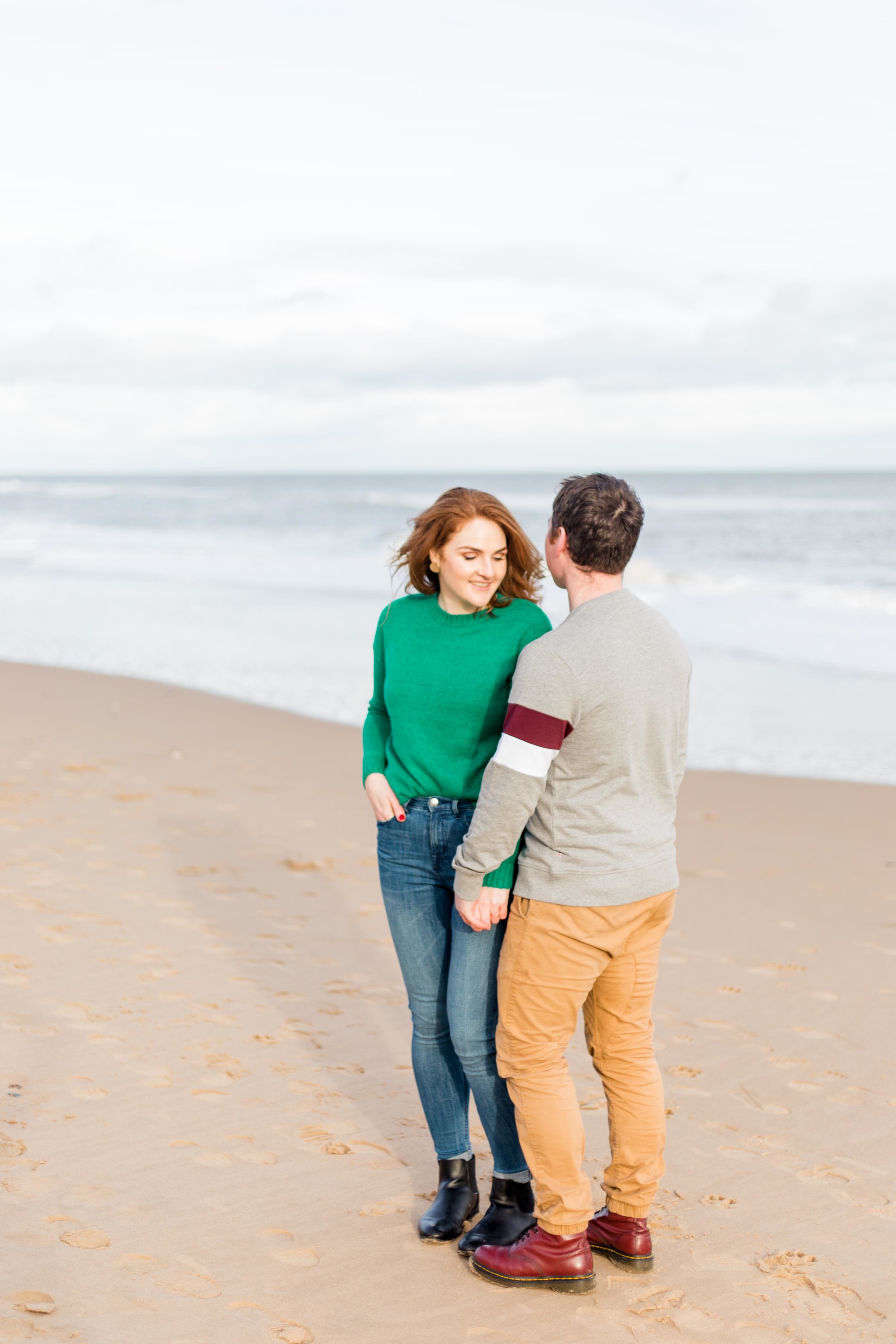 a couple is posing for a sunset photoshoot at brittas bay which is a sandy beach a woman wearing a green jumper with red hair and blue jeans a man wearing grey jumper with khaki chinos and brown boots