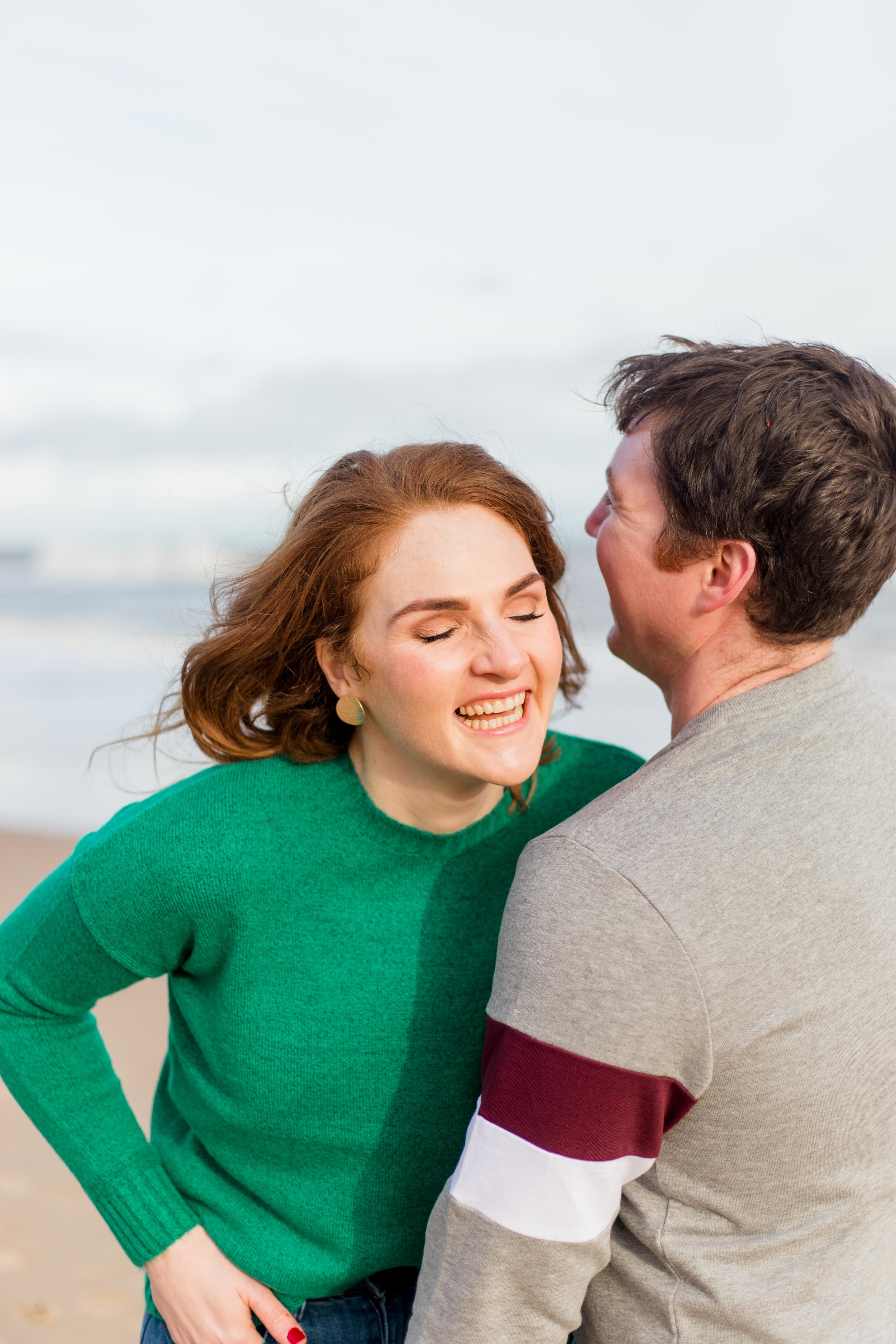 woman wearing a green jumper is being tickled by a man in grey jumper on a sandy beach posing for an engagement photoshoot at brittas bay in ireland