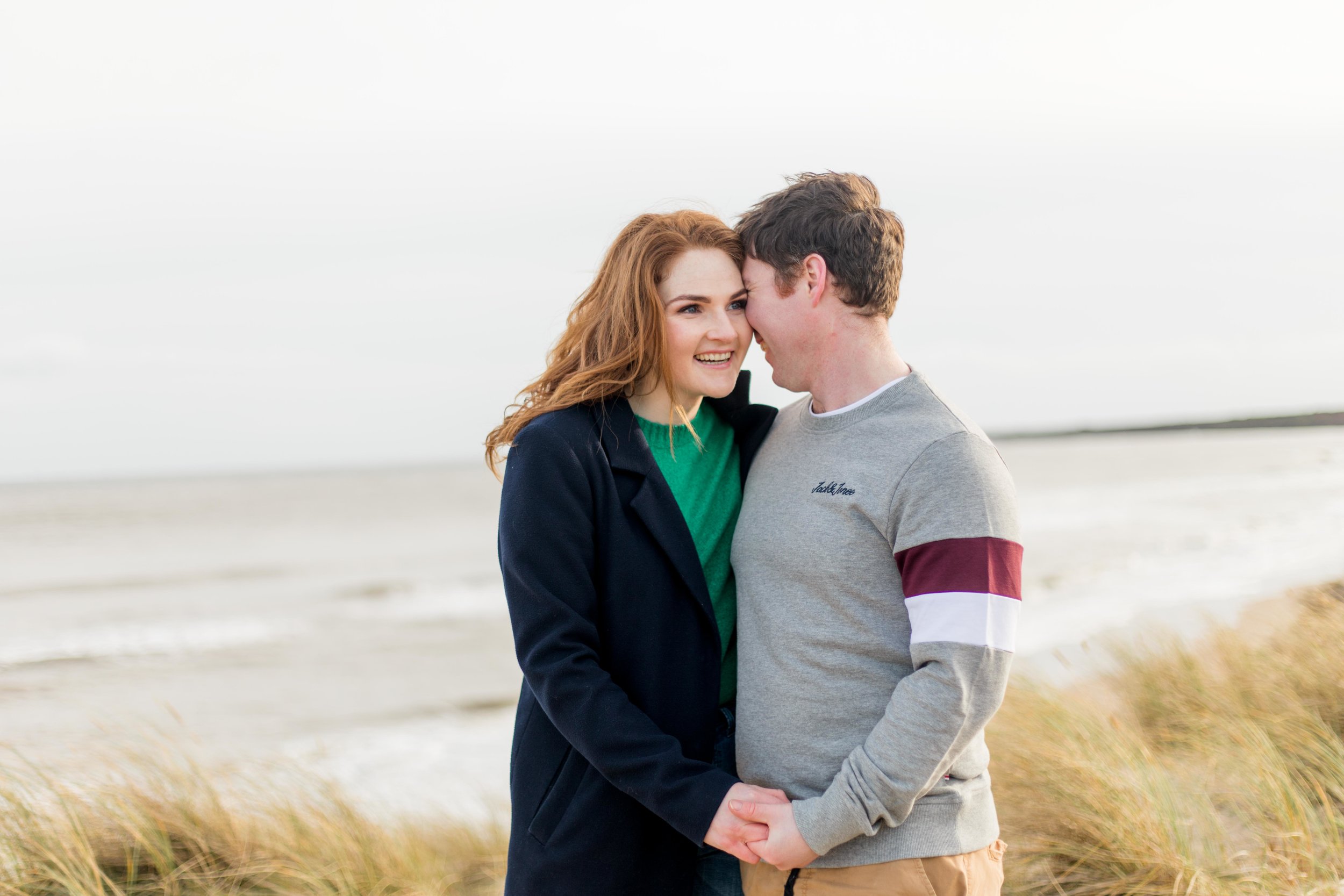 a couple posing for a photoshoot on a sandy beach a woman is dressed in green knit jumper and a black coat with red hair smiling at her fiancee in ireland