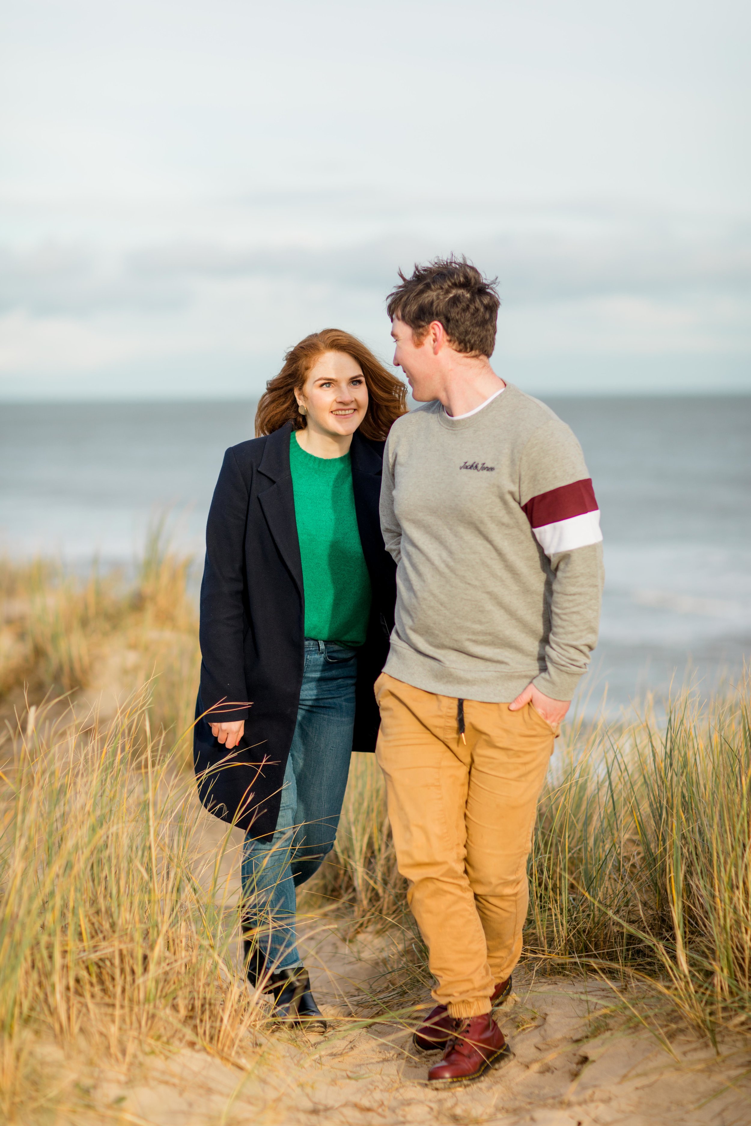 an engaged couple walking on a sandy beach in ireland brittas bay holding hands smiling looking at each other wearing a black knee length coat and khaki chinos