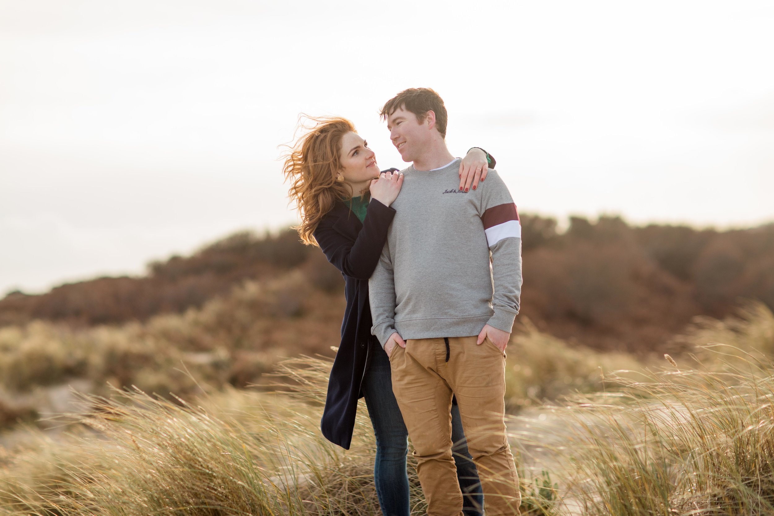a couple posing for their engagement session on a sandy dune in brittas bay during a sunset with wind blowing through her red hair while she is hugging her fiance