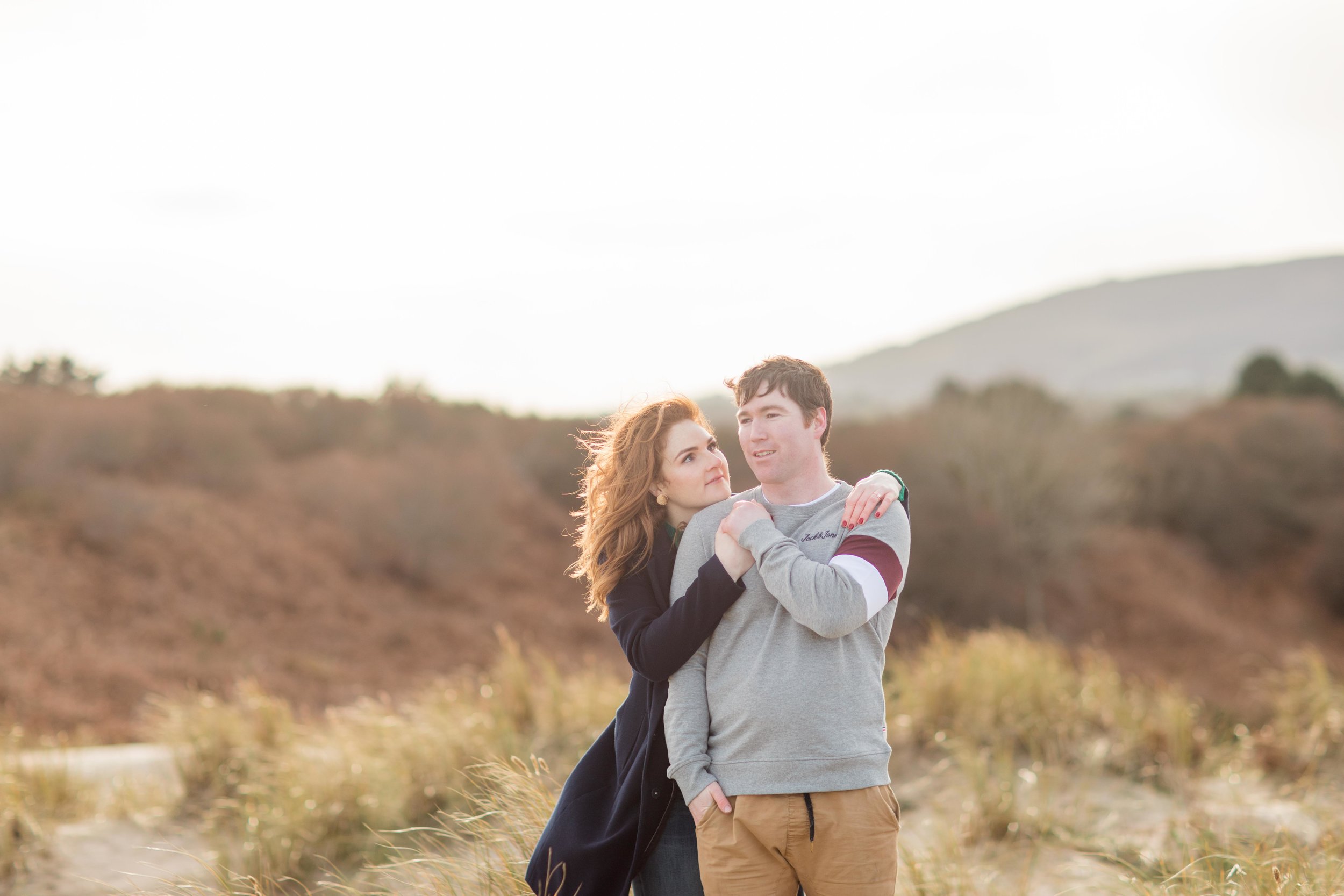 a sandy dune in brittas bay in ireland with a couple posing for a photoshoot and hilly background her hair red in a setting sun
