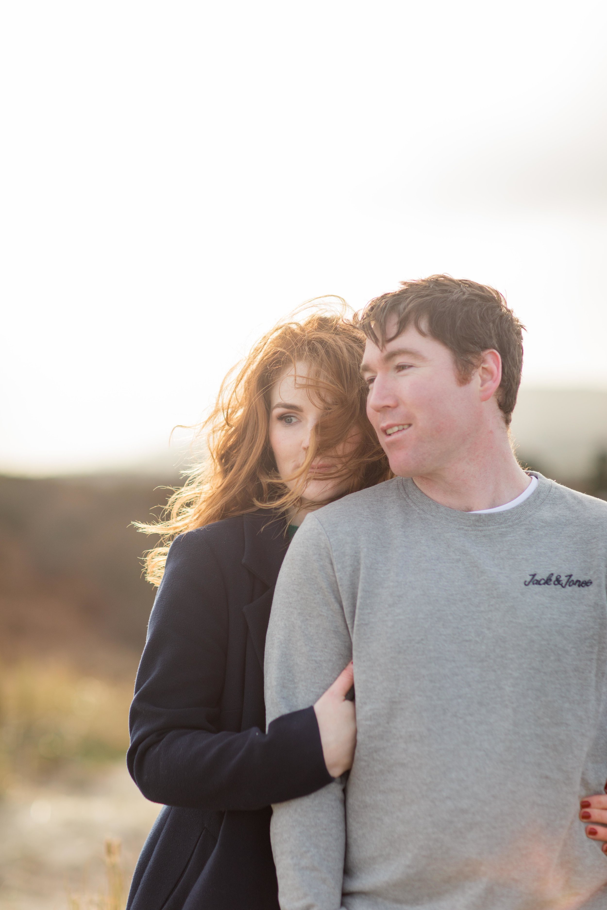 a couple posing for a photoshoot with her red hair caught in the wing wearing a black coat standing behind her fiance wearing a grey jumper in Ireland wicklow
