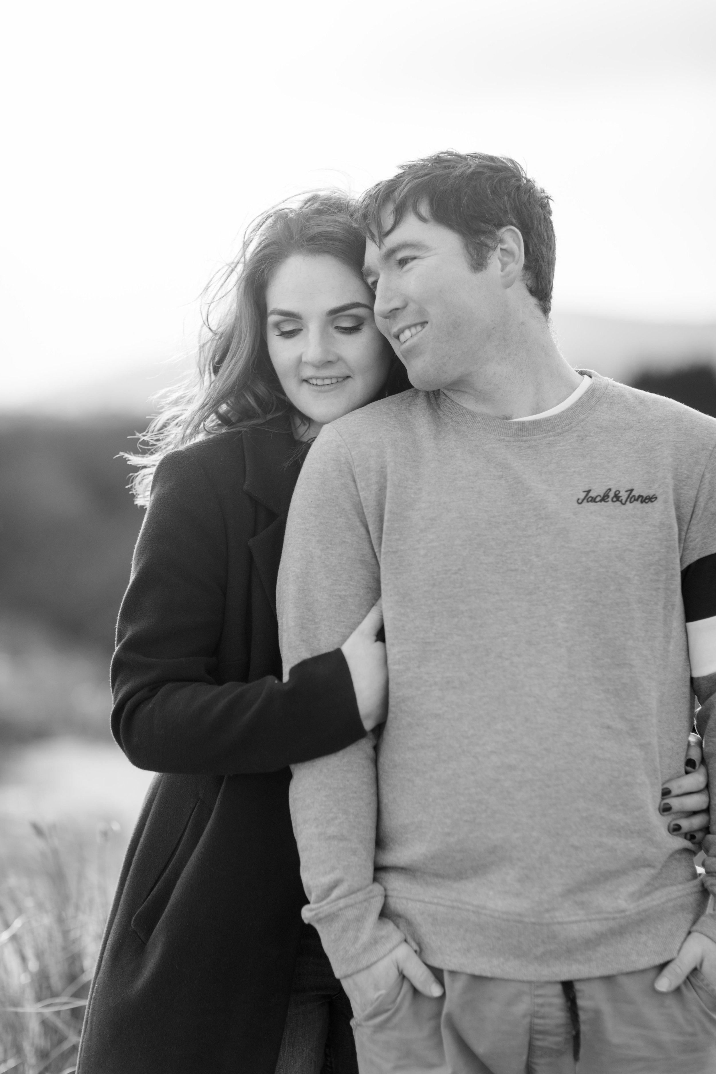 a black and white photo of a couple smiling she is standing behind her future husband wearing a black masculine coat