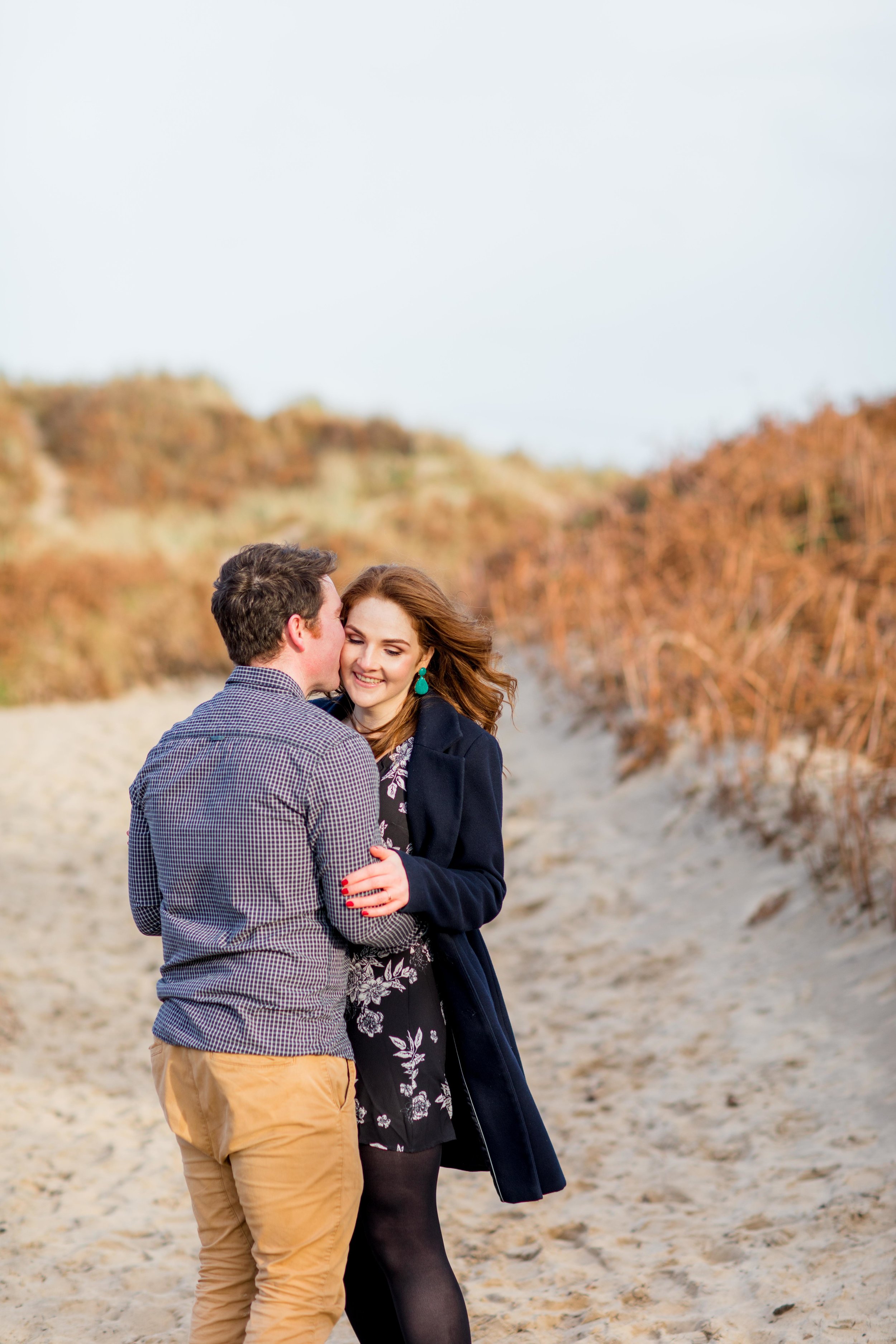 a couple posing for an engagement shoot wearing a black checkered shirt with khaki chinos and a knee length black dress with white flowers and green earings