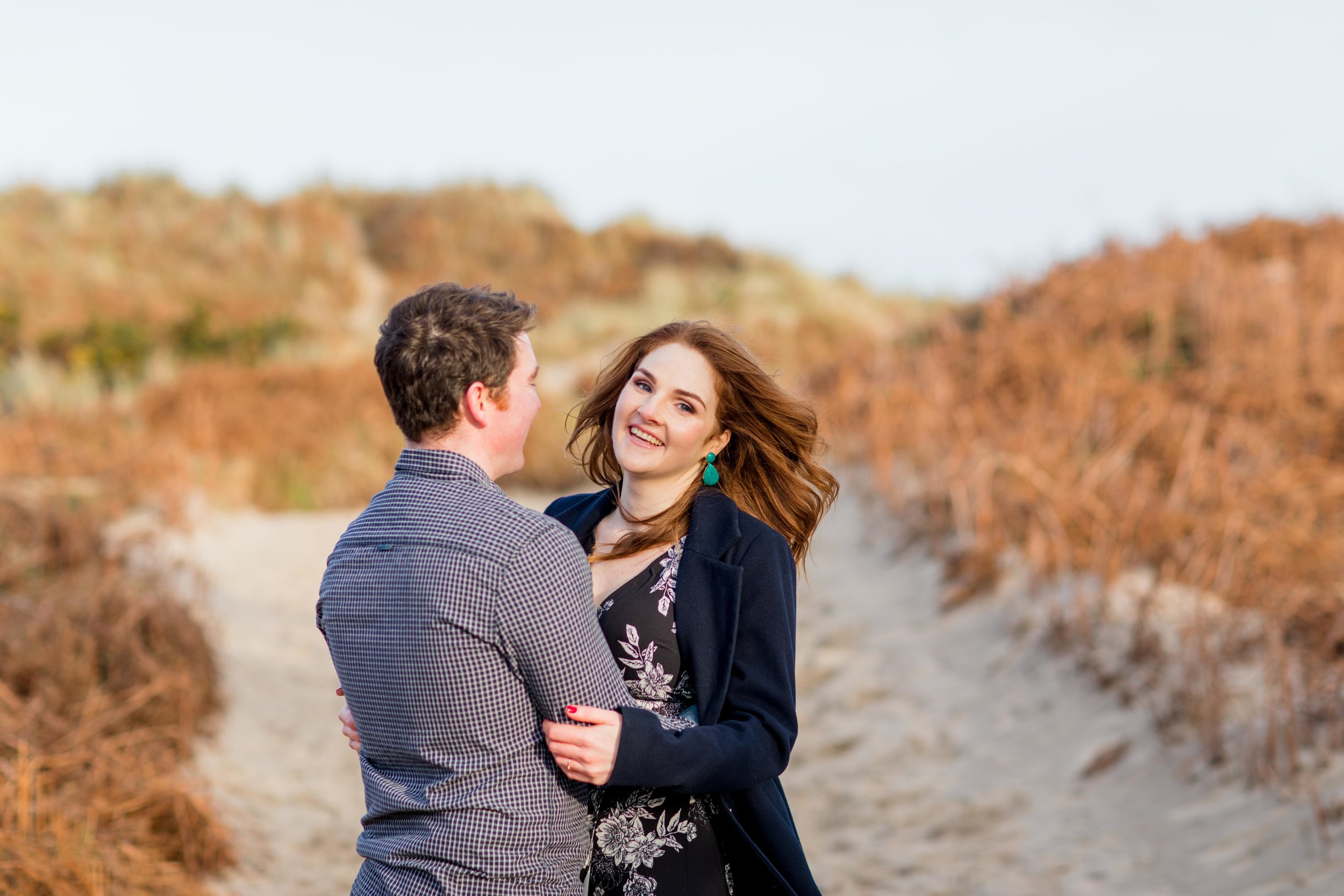 red haired woman wearing a black dress with white flowers and green earrings and a masculine coat with red nail varnish smiling at her future husband wearing a checkered shirt for an engagement shoot