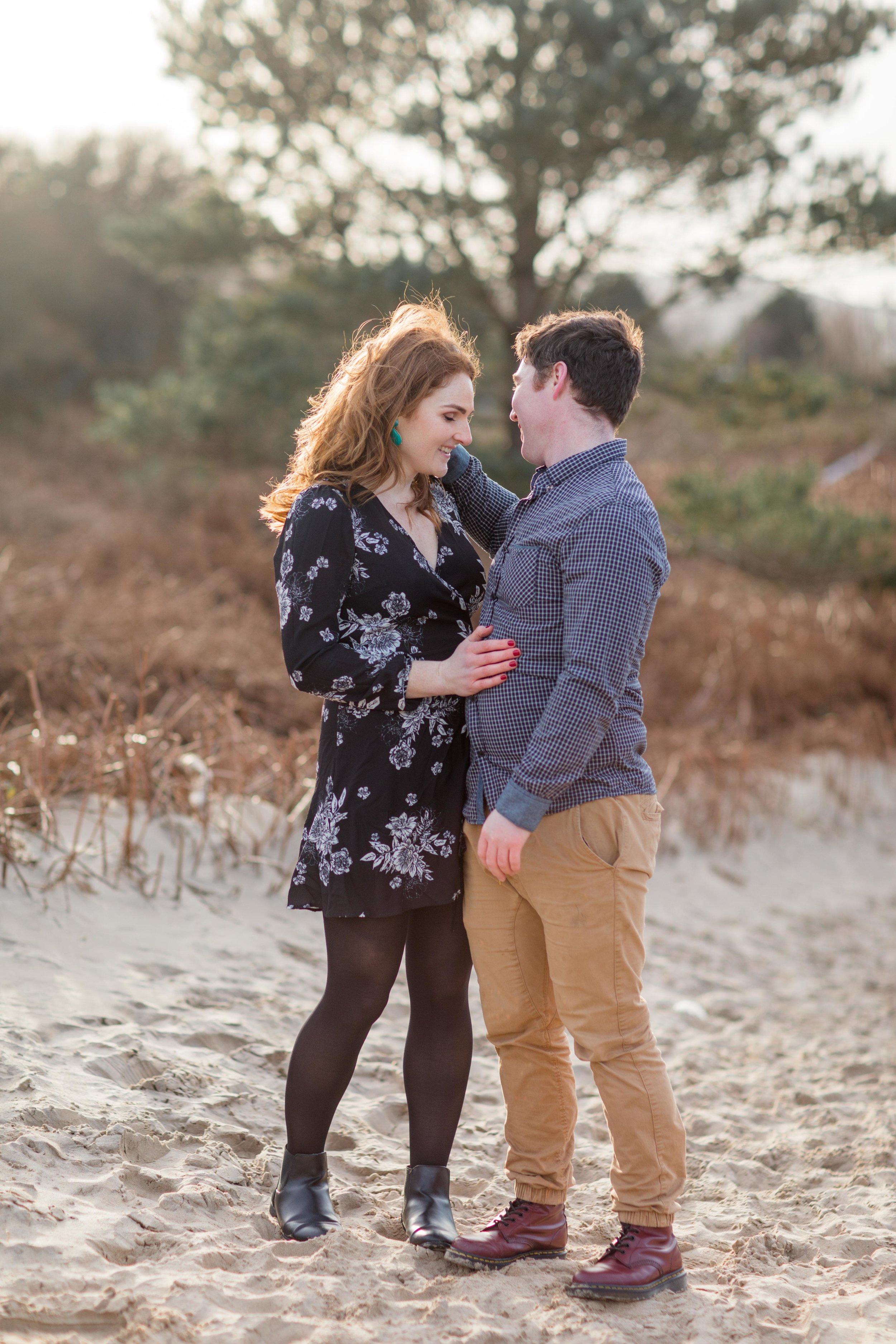 a future husband putting red hair behind his fiancees ear who is wearing a red nail varnish and a black dress with white flowers and ankle boots