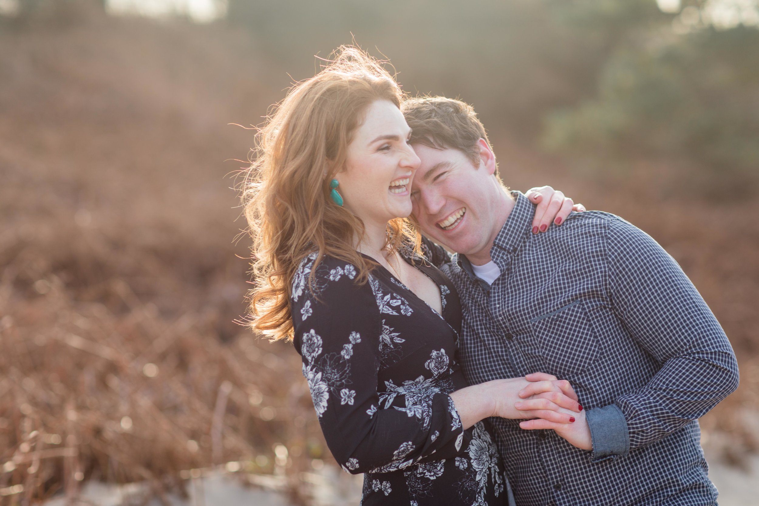 a couple holding hands and laughing a woman wearing black dress with white flowers and green earrings with red hair her fiance wearing black checkered shirt and a white t-shirt