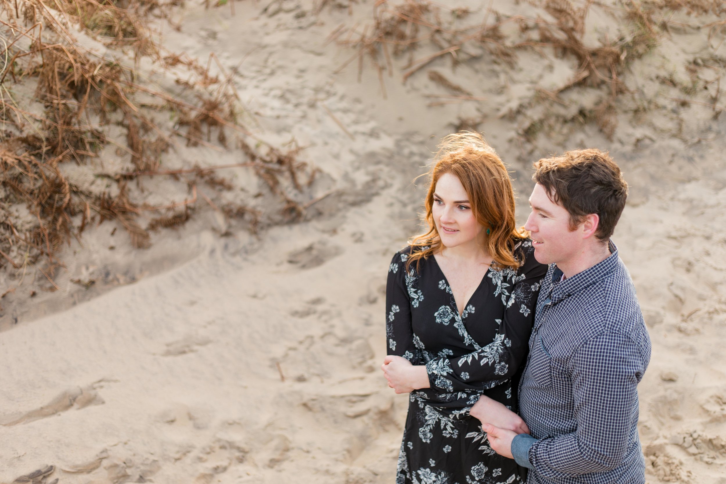 a red headed woman holding hands with her future husband posing for an engagement photoshoot with shoulder length hair wearing a knee length black dress with white flowers on a sandy beach in ireland