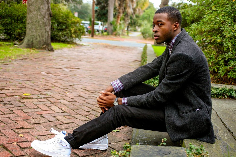 Menswear portrait of a man in a charcoal coat seated beside a brick path