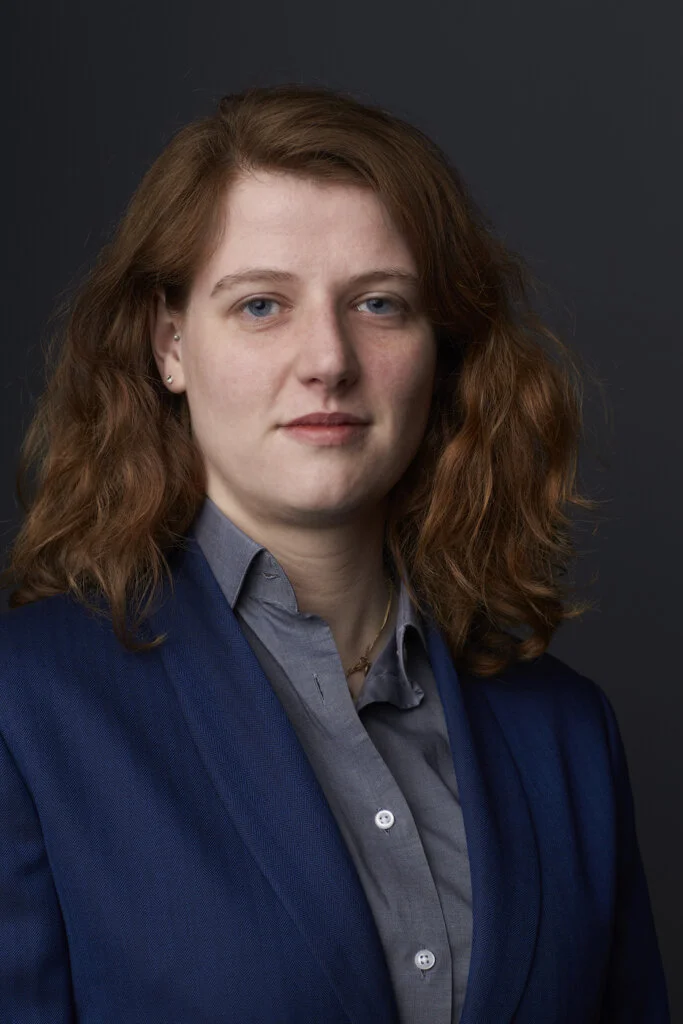 Portret van Amy, kleermaker en patroontekener bij De Oost Bespoke Academy, in een donkerblauw jasje op maat met grijs overhemd