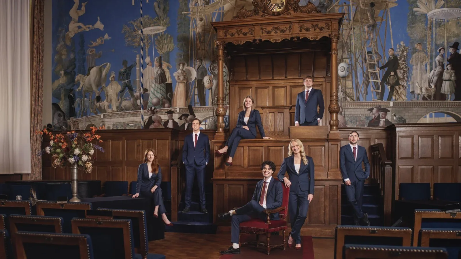 Studentenbestuur in donkerblauwe pakken op maat, dames en heren in dezelfde stof, gefotografeerd in een sierlijke hal