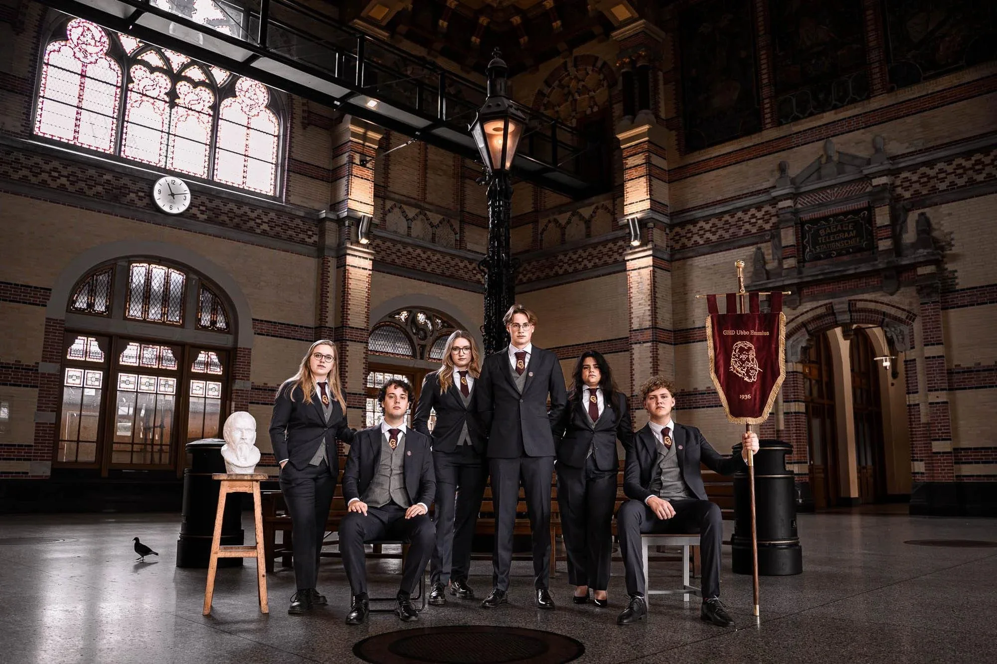 Gemengd studentenbestuur van zes personen in op maat gemaakte donkerblauwe pakken met bordeauxrode stropdassen, geposeerd in een historische bestuurszaal