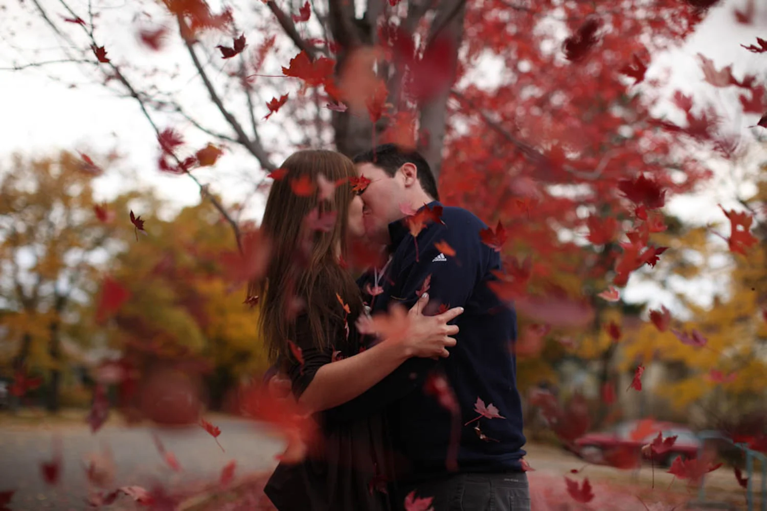 Red fall leaves falling around couple kissing during engagement portrait